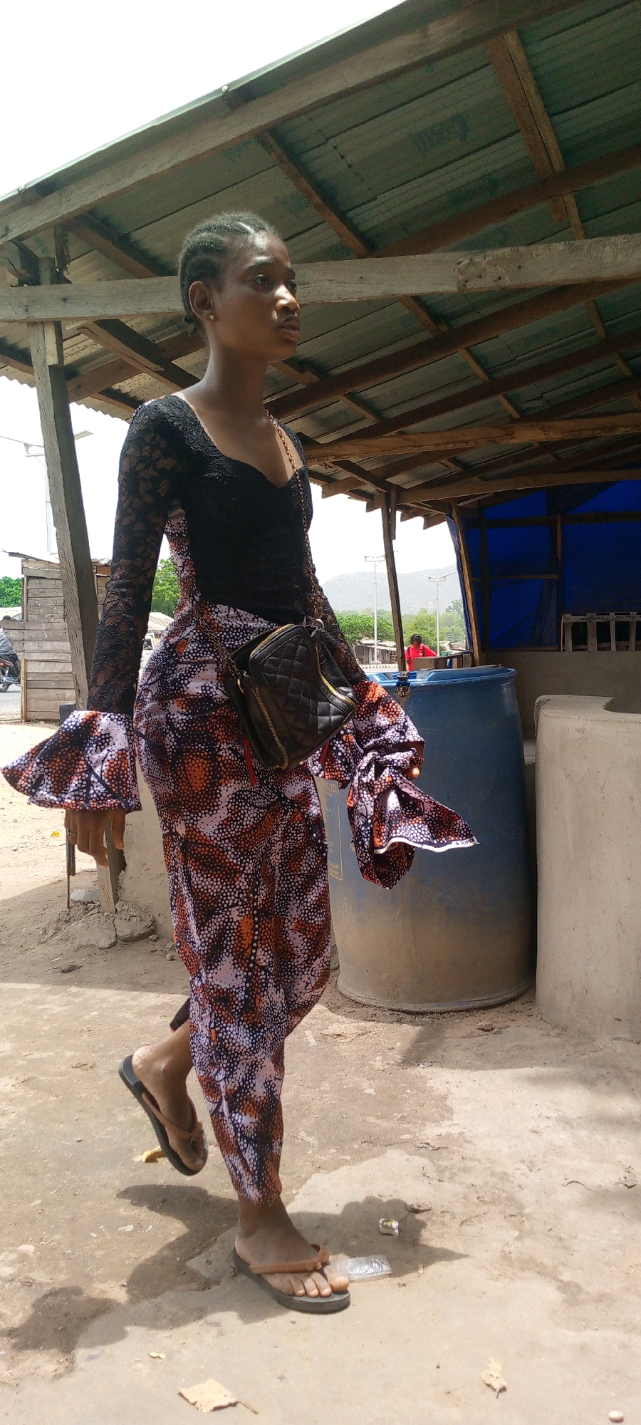 A woman is walking outdoors in Adankolo, Nigeria. She is wearing a black lace top with long, flared sleeves and a long, patterned skirt. She also has on a small, quilted black purse slung over her shoulder. She is wearing flip-flops. The ground is dirt and sand, and there is a crude structure with a tin roof and wooden supports nearby. There is a large blue barrel and some concrete structures behind her. In the background, there are other buildings, a street, and a tree-covered hill.