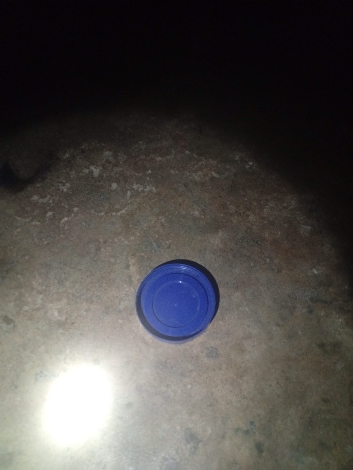 A blue plastic lid is on the ground in Jalingo, Nigeria. The lid is circular with a raised rim. The ground is a rough, textured surface, possibly concrete or asphalt, with a light-colored, speckled pattern. A bright white light source illuminates the lower left portion of the image, creating a strong glare. The upper portion of the image is dark and indistinct. The scene appears to be outdoors at night.
