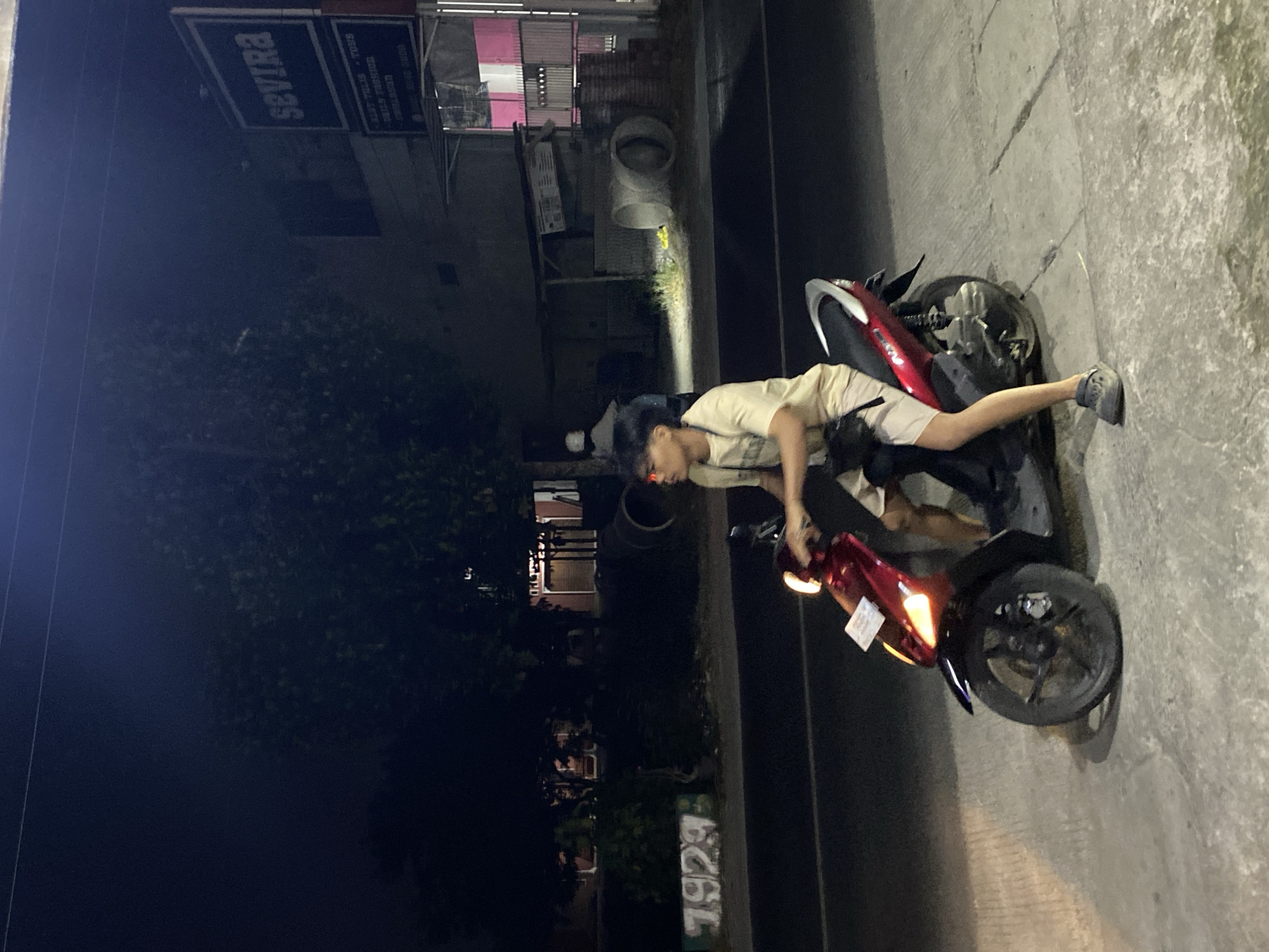 A young man wearing a light-colored t-shirt and shorts is sitting on a red motorcycle at night on a street in Kasihan, Indonesia. The motorcycle's headlight is on, illuminating the area in front of it. The man is looking to his left. In the background, there are trees, buildings, and a sign that reads "SEVIRA". The street appears to be paved. There are some cylindrical concrete objects on the right side of the image, and graffiti that says "1929" on the left. The scene suggests an urban or suburban environment at night.