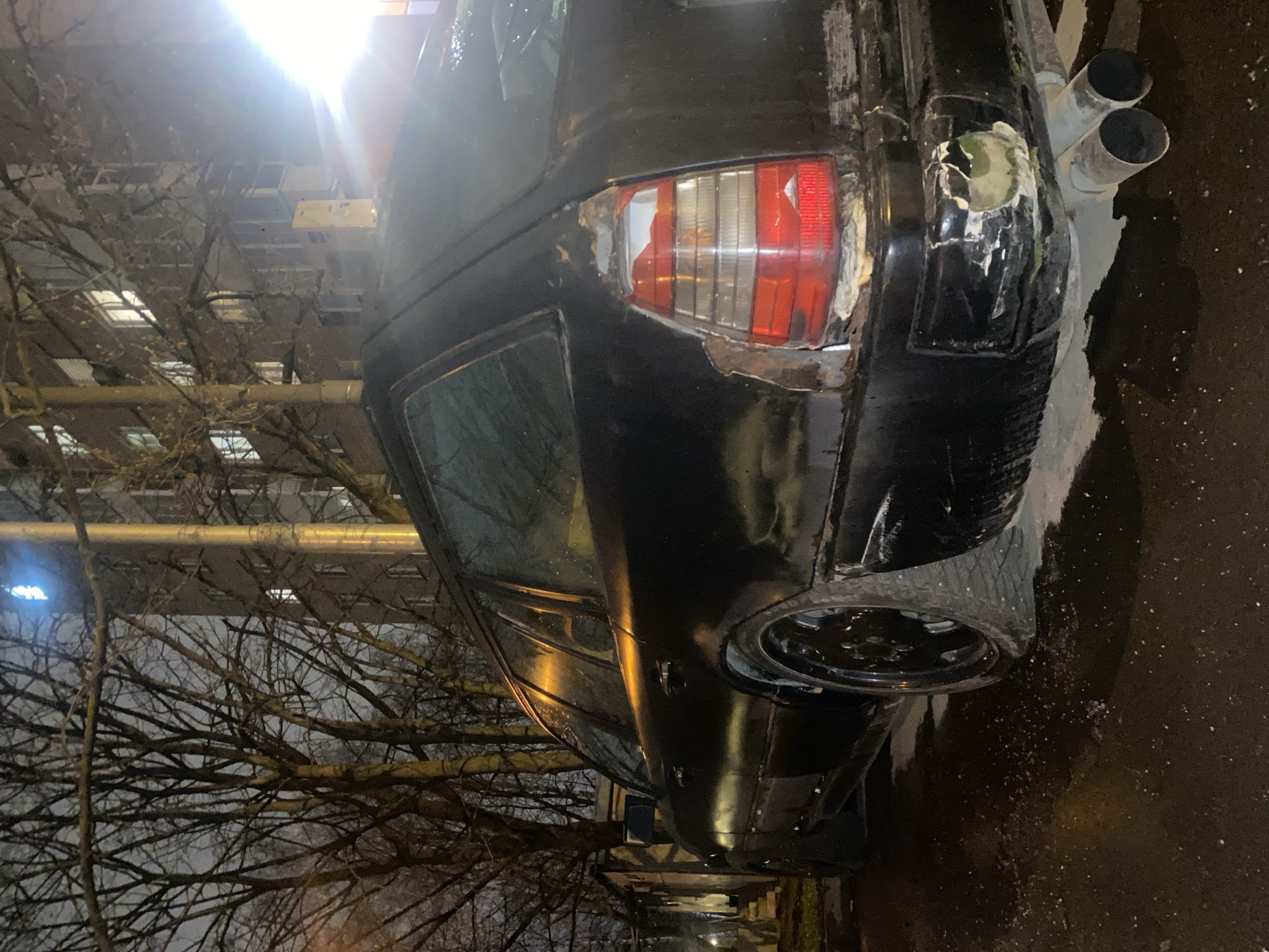 The image depicts the rear end of a black car, possibly a Mercedes-Benz W124, parked on a street at night. The car has visible damage to the rear panel near the right tail light and a non-factory exhaust system with dual pipes. The vehicle appears to be in disrepair or a project car.
The surrounding environment suggests an urban setting. A bare tree is visible in the foreground with a building illuminated in the background, indicating that the scene is potentially located in a residential or commercial area within a city. The presence of moisture on the pavement indicates recent or current wet conditions.
Given the urban environment and architecture, it's plausible the photograph was taken in Moscow, Russia. However, definite confirmation would require additional details.