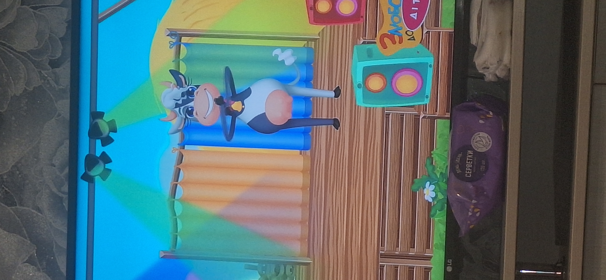 A cartoon cow with black and white spots, a bell around its neck, and wearing a pink udder is standing on a wooden stage. The cow is smiling and has its arms outstretched. Behind the cow are three curtains of different colors: green, orange, and blue. There are two spotlights shining down from above. To the right of the cow are two speakers, one teal and one red, and a sign with Cyrillic text. In front of the stage, there is a small white flower with green leaves. Below the stage, a purple package of wet wipes with Cyrillic text is visible. The overall scene appears to be from a children's game or animation. There is no indication of the location being in Bila Tserkva, Ukraine.