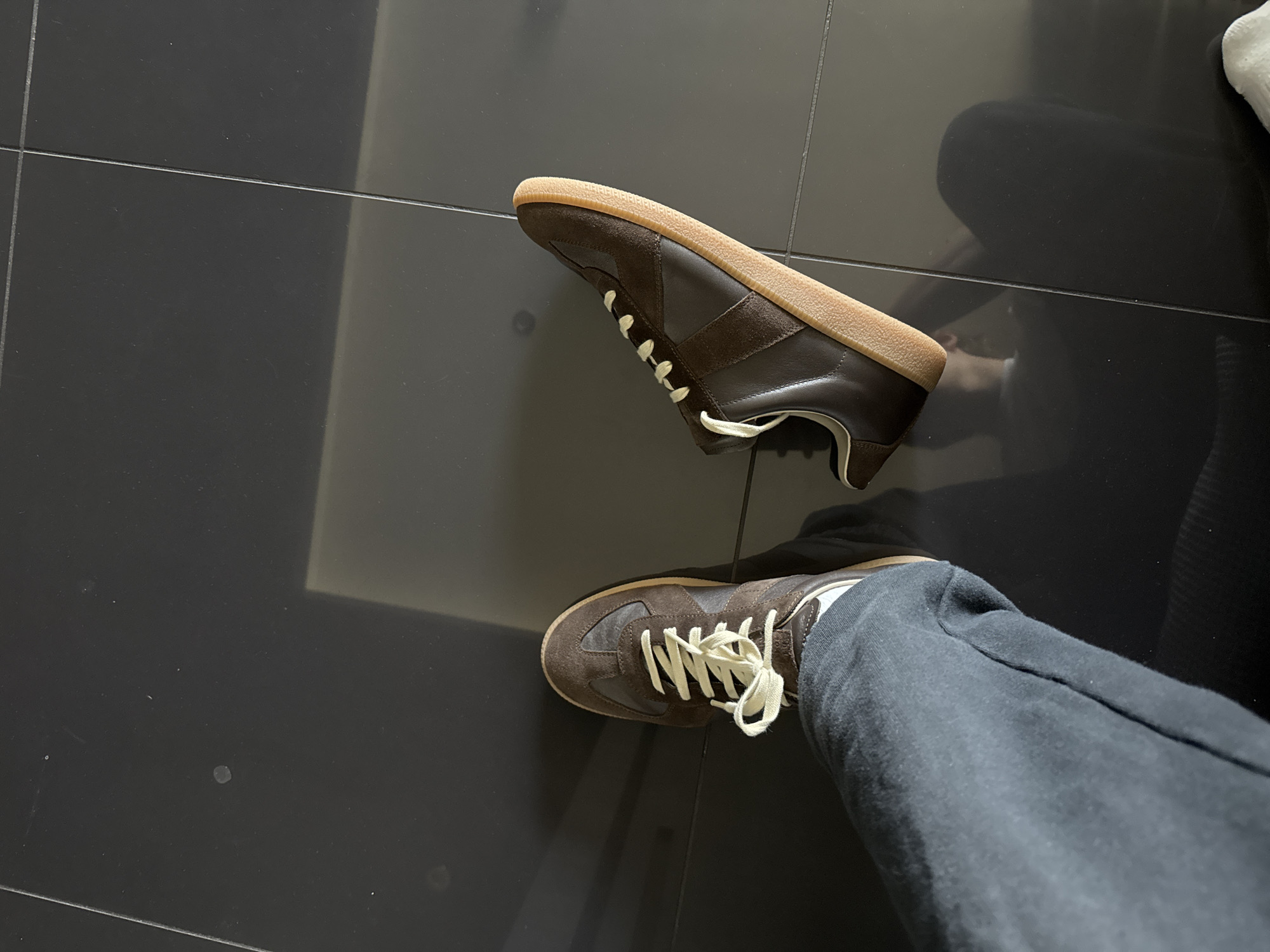 The image shows a person wearing a brown and tan sneaker on a dark, tiled floor. Another identical sneaker lies on the floor next to the person's leg. The person is wearing dark gray sweatpants. The location is not specified in the image and cannot be determined from the available information. No actions or interactions are visible.