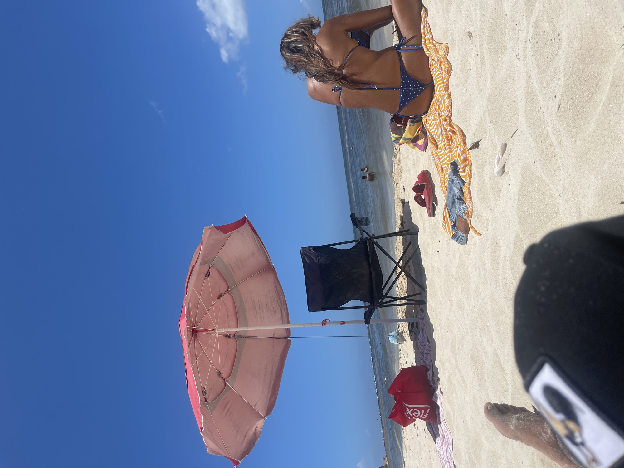 The image shows a beach scene at Playa del Carmen, Mexico, on a sunny day. A woman wearing a blue polka dot bikini is sitting on a patterned towel. Nearby are a pair of red sandals, a pair of denim shorts, and a yellow patterned cloth. A black folding chair and a red shopping bag from "Flex" are also present. An open beach umbrella with a red and white canopy stands near the chair. The clear blue sky is visible above, and the ocean stretches to the horizon with a few people visible in the water. A person's foot and part of a black baseball cap is in the foreground.