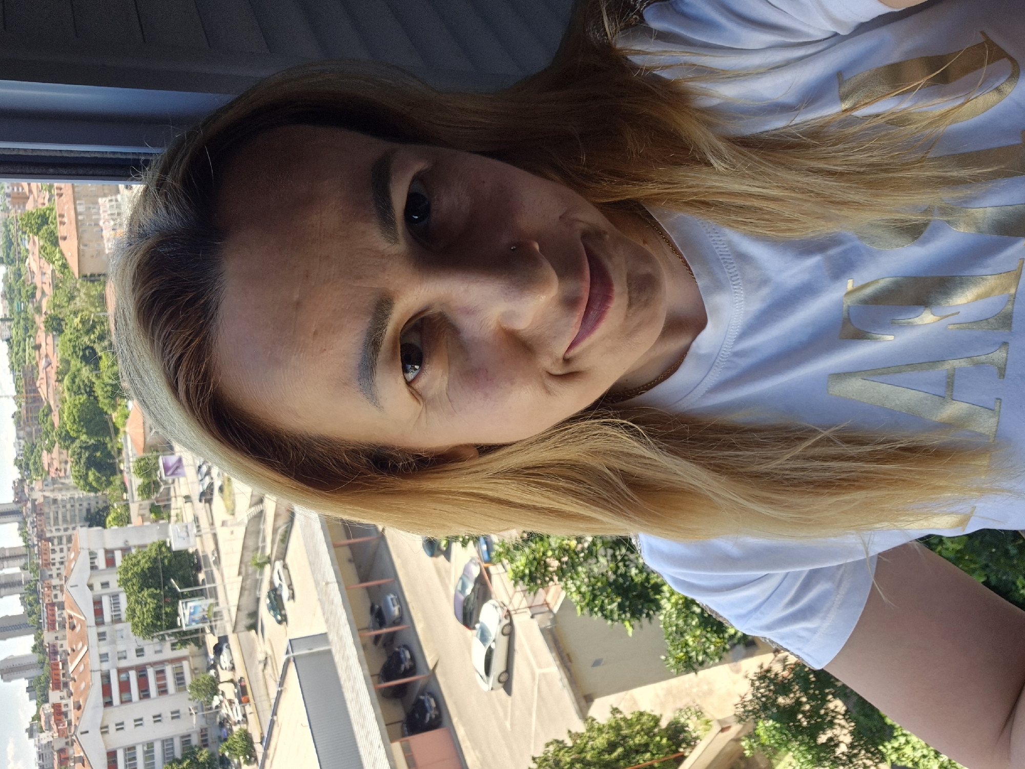 A medium shot portrait of a woman with blonde hair and a white t-shirt is captured from the chest up. She is standing on a balcony or elevated position, looking into the camera with a slight smile. The background reveals a city landscape with buildings of varying heights, some with orange tiled roofs, and a street below with parked cars. Green trees are visible in the mid-ground and foreground. The lighting suggests it is daytime.