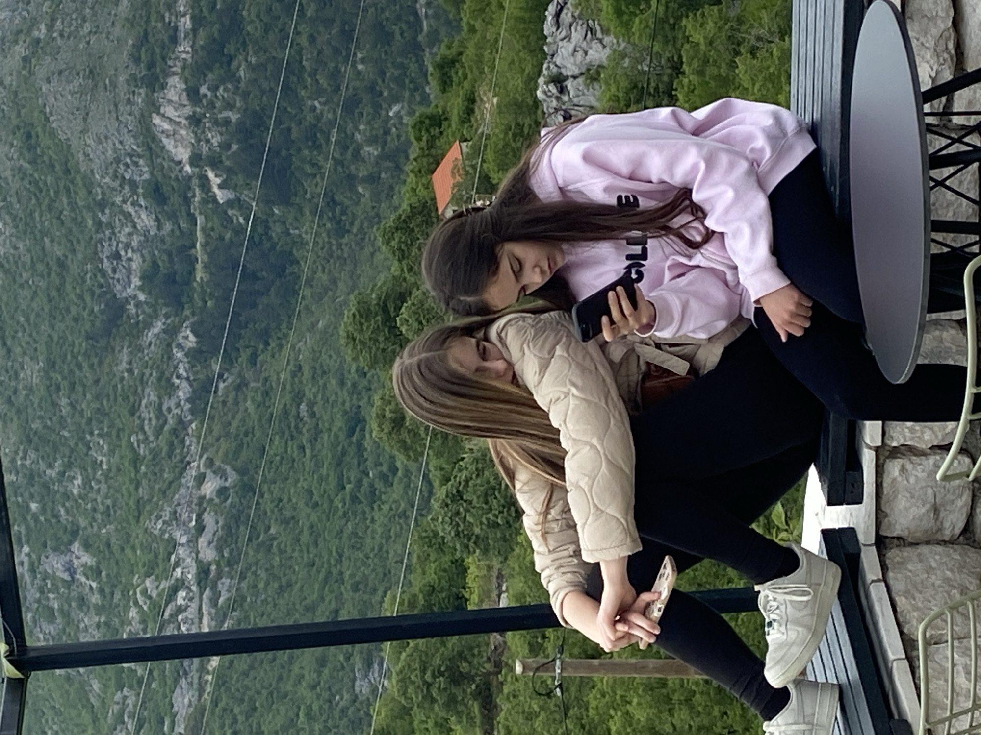 Two young women are sitting on a ledge in Lipa Dobrska, Montenegro, overlooking a mountainous, forested landscape. One woman is wearing a pink sweatshirt and black leggings, looking at a smartphone. The other woman is wearing a quilted beige jacket and black leggings, holding a different smartphone. They are sitting near a small metal table and a metal fence. The backdrop shows steep rocky terrain covered in dense green trees and vegetation. Overhead power lines are visible in the background.