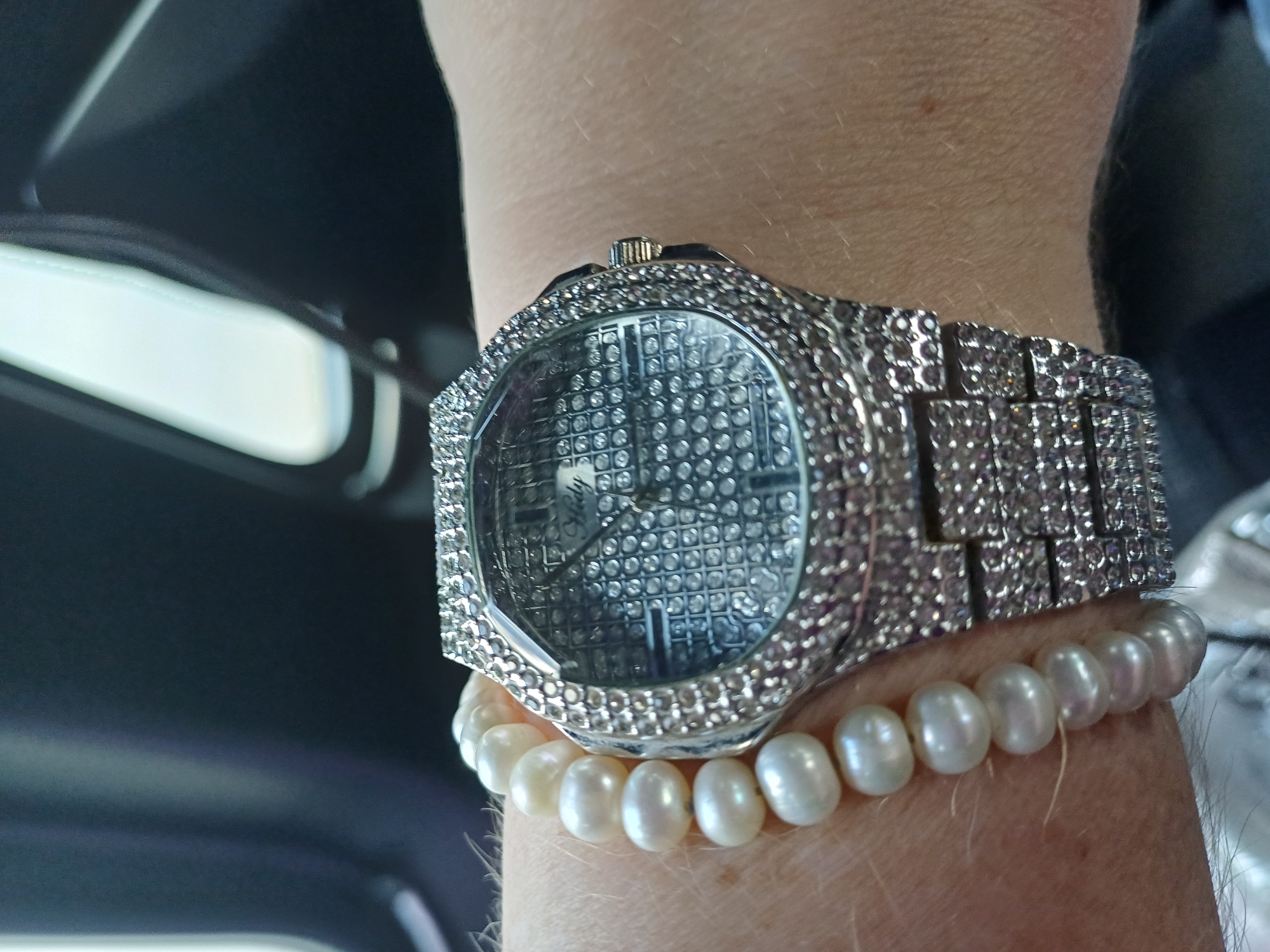 The image shows a close-up of a person's wrist adorned with a pearl bracelet and a diamond-encrusted watch. The watch has a metallic band and a faceted bezel. The dial of the watch appears to be covered in small, reflective square patterns, and a brand name, "Hedy," is visible in the center. The background is dark and out of focus, suggesting an indoor or vehicle setting. There is no indication of the location within the city of Maiaky, Ukraine.