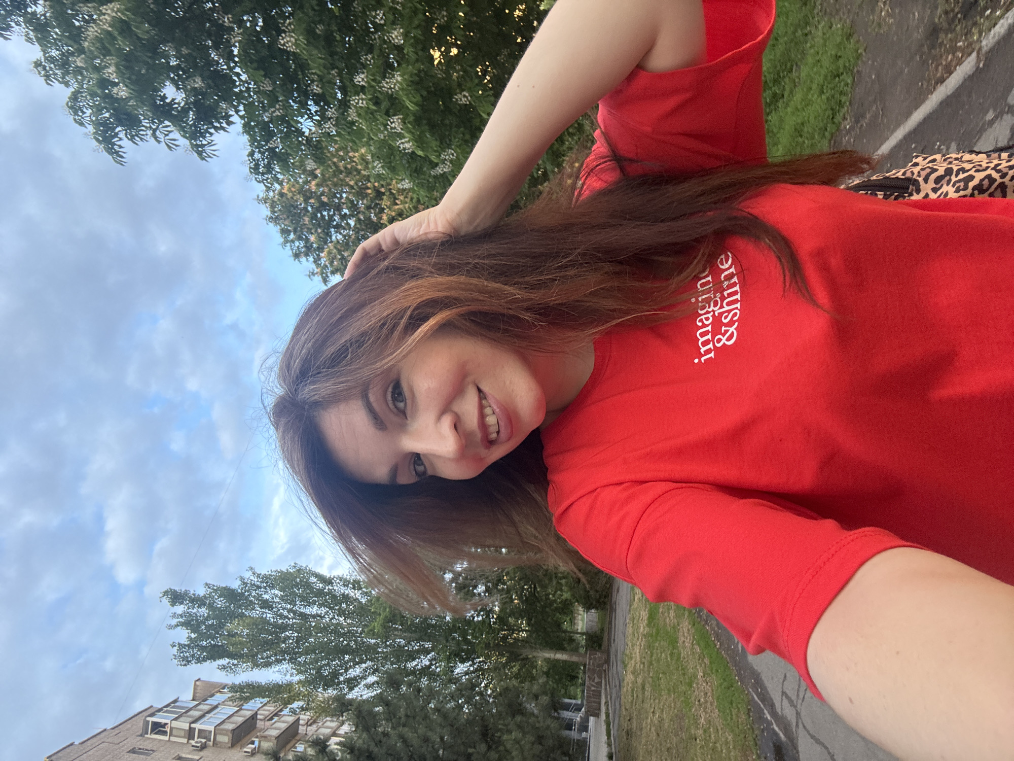 A smiling woman is taking a selfie outdoors. She is wearing a red t-shirt with the words "imagine & shine" printed on it. Her right hand is raised and appears to be touching her hair. In the background are trees and a cloudy sky. Based on the architecture of the building visible on the left side of the image, this photo was taken in the city of Kryvyi Rih, Ukraine.