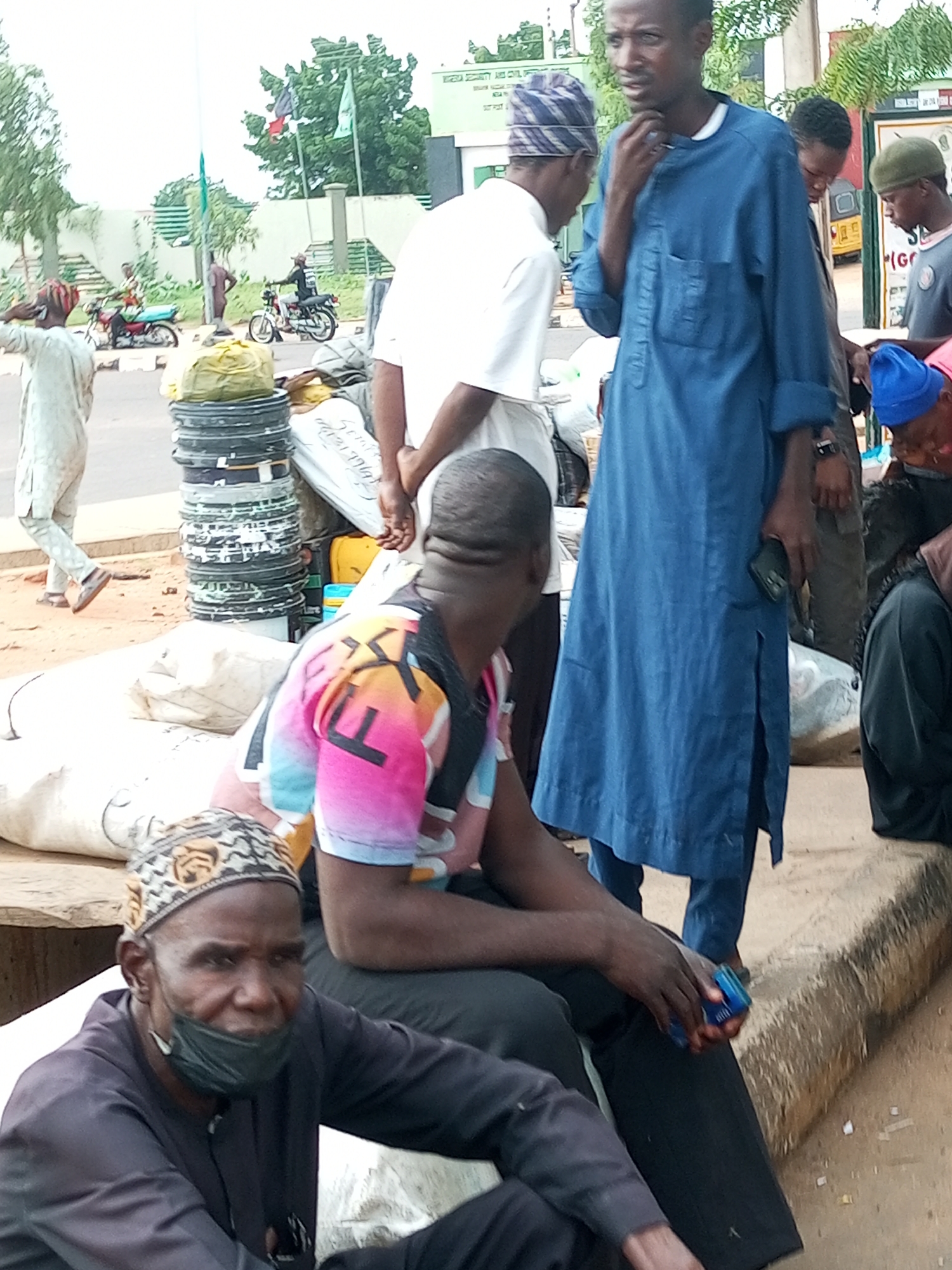 The image depicts a street scene in Gombe, Nigeria, likely near a market or transport hub. In the foreground, a man wearing a dark traditional outfit and a patterned cap is seated and looking towards the camera. He has a black face mask pulled down to his chin. To his right, another individual in a bright, colorful shirt with "FX" printed on it is also seated. Behind them, a man in a blue traditional robe stands, holding a blue object, possibly a phone or a small container, in his right hand. A stack of black, circular objects, resembling used tires or industrial rings, is prominent in the mid-ground, alongside large white sacks and a yellow container. Several other people are visible in the background, including some on motorcycles. The environment appears to be dusty and informal, with a curb running along the right side of the frame. The overall impression is of a busy, everyday scene in an urban Nigerian setting.