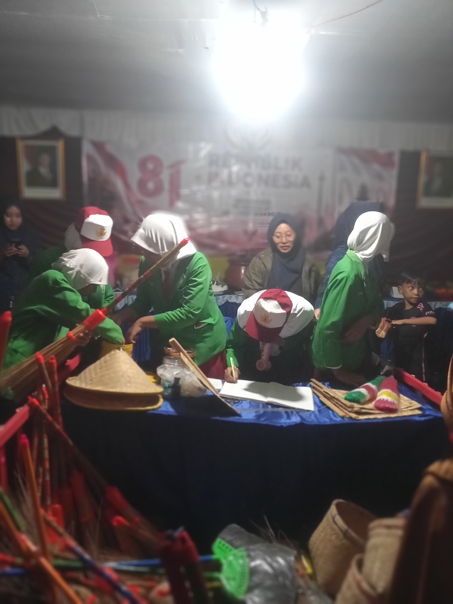 The image depicts a dimly lit indoor scene, possibly a market or event stall, in Pule, Indonesia. Several people, predominantly young women wearing green jackets and white headscarves, are gathered around a blue-covered table. Some are actively engaged in tasks, such as one individual writing in a notebook and others holding what appear to be brooms or sticks with red tips. A woman wearing glasses and a dark headscarf is also present, looking towards the activity. A young child in a black t-shirt stands to the right of the table, observing the scene. The background features a banner with large numbers "81" and text that appears to be in Indonesian. Various objects are on display or in use, including woven conical hats, a glass jar, what looks like a bundle of dried grass or reeds, and some colorful items resembling candy or small toys. The overall atmosphere is informal and suggests a community gathering or commercial activity.