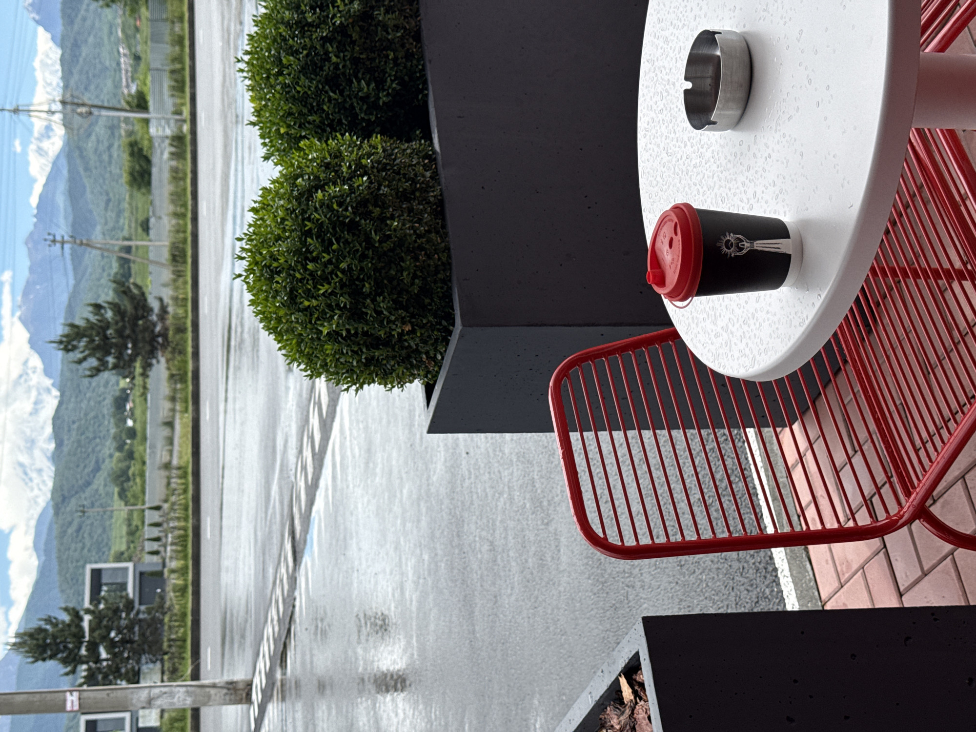 The image depicts an outdoor seating area at a cafe located in Ir, Russia, with a backdrop of mountains. In the foreground, a white, round table is wet with raindrops, holding a black paper coffee cup with a red lid and a silver ashtray. A red slatted metal chair is partially visible to the left of the table. The ground is paved with red bricks, and behind the table are large, dark gray planters containing trimmed green bushes. Beyond the planters, a wet asphalt road with white markings is visible, suggesting recent rain. In the middle ground, a modern building with dark facades and large windows stands among trees. The background features rolling green hills and snow-capped mountains under a cloudy sky, characteristic of the mountainous region around Ir.