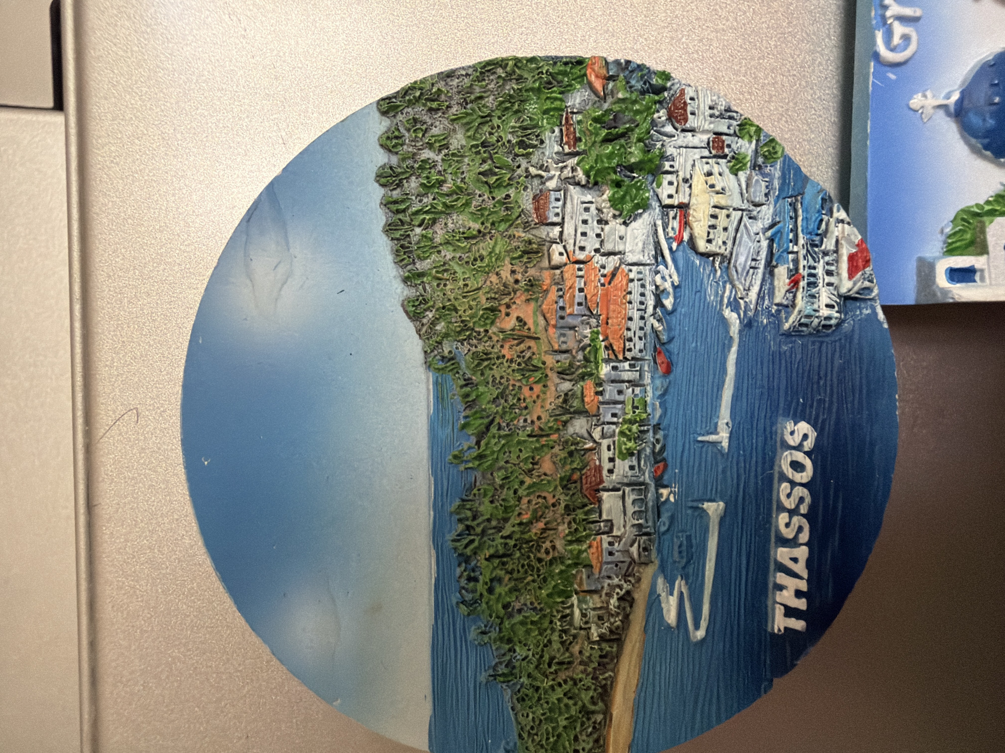 This is a close-up, eye-level shot of a circular souvenir magnet featuring a miniature embossed depiction of the Greek island of Thassos. The magnet shows a coastal scene with white buildings with orange roofs clustered on a hillside covered in green vegetation. The buildings are situated along a blue, textured body of water that represents the sea, where several small boats are docked. In the foreground, the word "THASSOS" is embossed in white capital letters on a dark blue background. The magnet is placed on a textured, light-colored surface, likely a refrigerator or a similar metallic appliance. The lighting is slightly uneven, casting some shadows and highlighting the three-dimensional details of the magnet. There are no people or animals present in the image. The magnet is partially obscured by another souvenir magnet in the lower right corner, which shows a blue dome and the letters "GY". This image is not taken in Pazardzhik, Bulgaria, as it depicts a scene from Thassos, Greece.