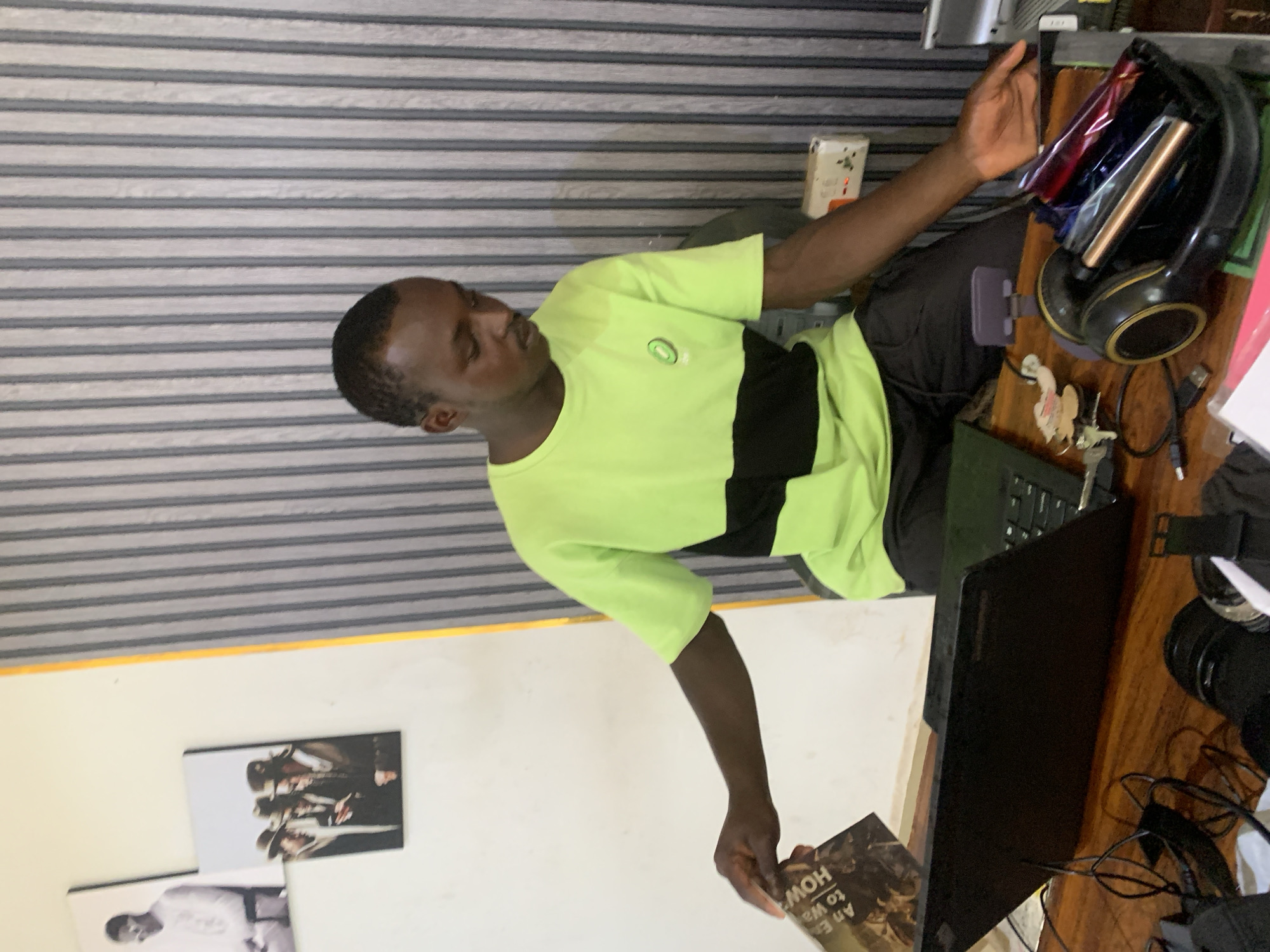 A man is seated at a desk, appearing to be working or studying. He is wearing a two-toned green and black t-shirt. On the desk are a laptop, headphones, keys, and various other electronic items. He is holding a book in his left hand.
Photographs are visible on the wall behind him. The wall has a striped pattern at the top. The location is plausibly Jos, Nigeria, based on the individual's appearance and general environment, but there are no uniquely identifying features in the image to definitively confirm it.