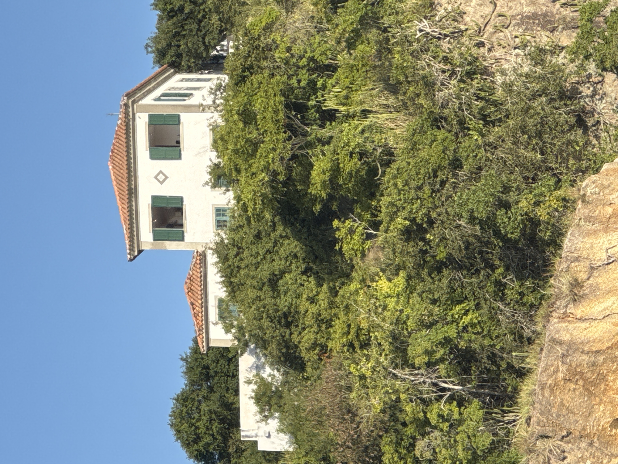 The image shows a white, two-story house with a red-tiled roof, perched on a hill covered in dense green foliage. The house has several windows with green shutters. One window on the second floor is open, revealing a glimpse of the interior. Another smaller building with a similar roof is visible to the left. The foreground consists of a rocky, textured surface with sparse vegetation. The sky is a clear, vibrant blue. This house is likely located in the city of Niterói, Brazil, given its hilly terrain and lush vegetation characteristic of coastal regions in Rio de Janeiro state. The scene depicts a peaceful, secluded dwelling surrounded by nature.
