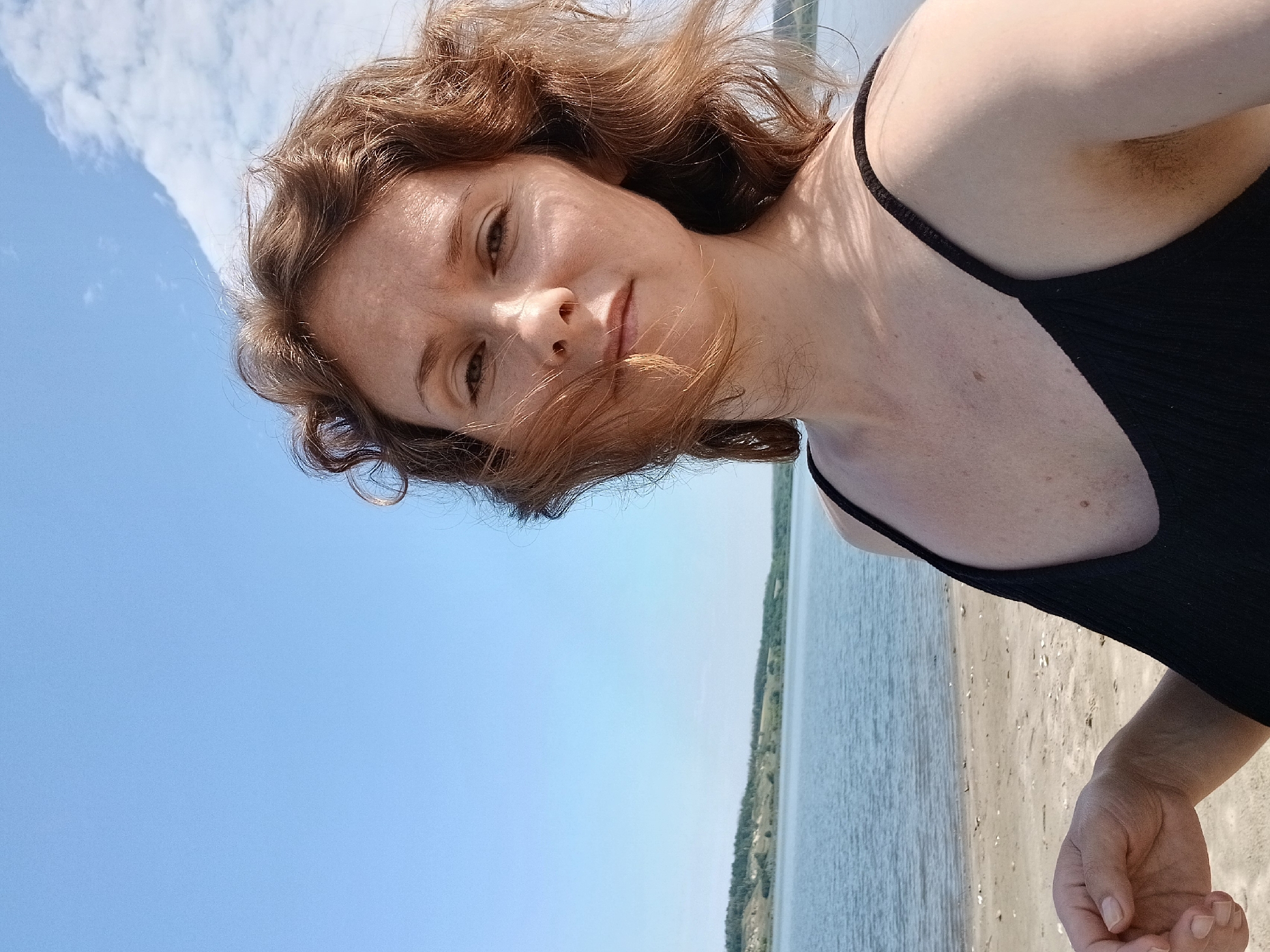 A woman with wavy brown hair is taking a selfie on a sunny day at the shore of a body of water, possibly the Don River in Maslova Pristan', Russia. She is wearing a black tank top. The background shows a sandy beach, calm blue water, and a green, rolling landscape under a clear blue sky with scattered white clouds.