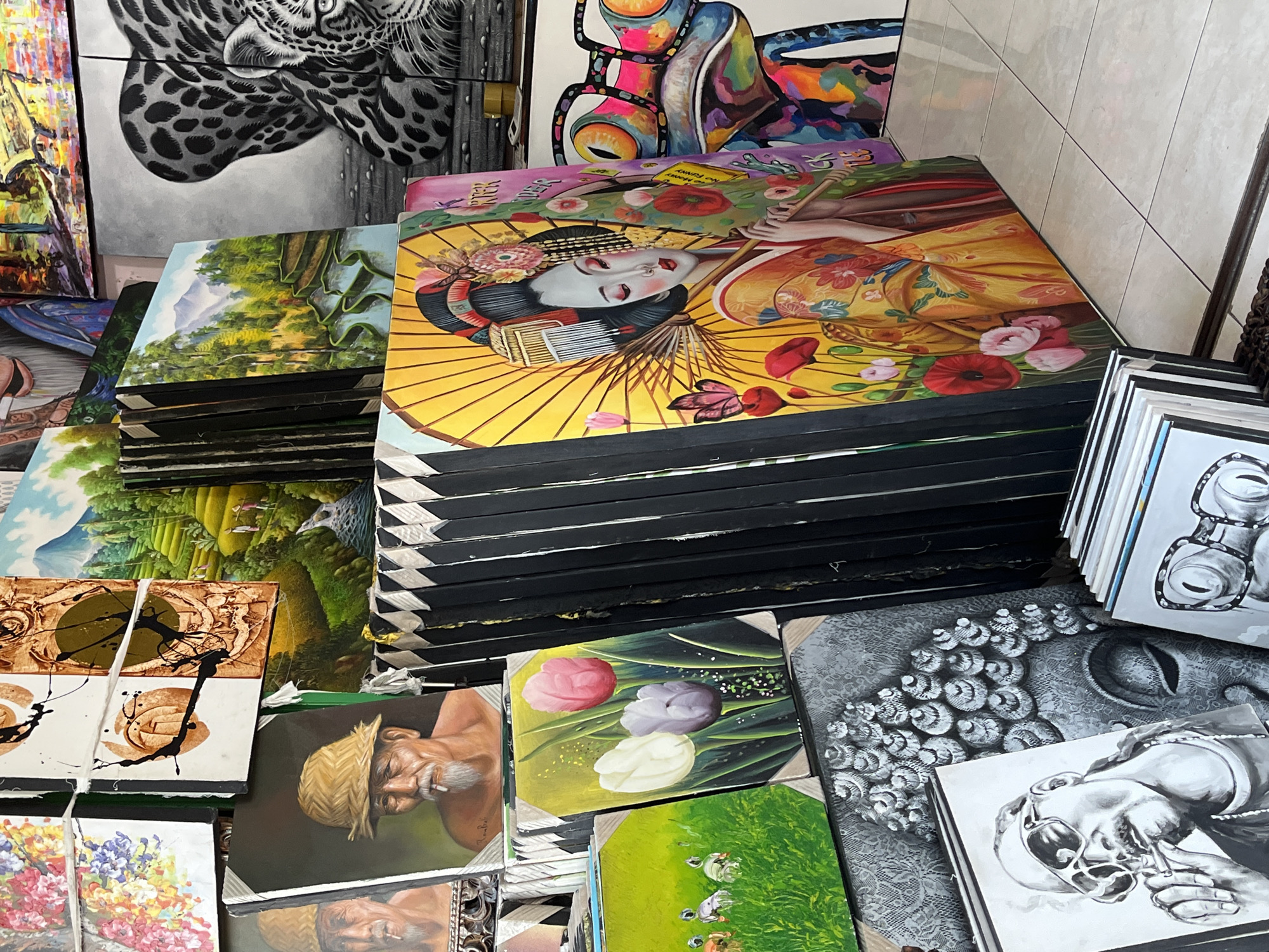 The image depicts a collection of stacked paintings in a shop or market setting in an unknown city in Indonesia. The paintings cover a variety of subjects, including landscapes, portraits, abstract art, and a depiction of a geisha. The paintings are stacked haphazardly, with some leaning against others and some arranged in neat piles. The background includes partial views of other paintings, including a black and white image of a jaguar or leopard. The setting appears to be a small, cluttered retail space with tile walls visible in the background.