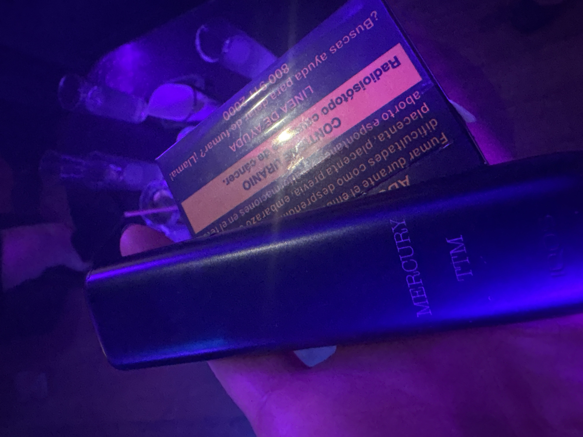 A hand holds a black Mercury TTM vaporizer next to a cigarette pack labeled “Radioisotopo.” The cigarette pack includes text in Spanish stating that smoking during pregnancy can cause complications and that the product contains Uranium. An assistance line number for quitting smoking is listed as 800-911-2000. The scene is illuminated with a blue/purple light.