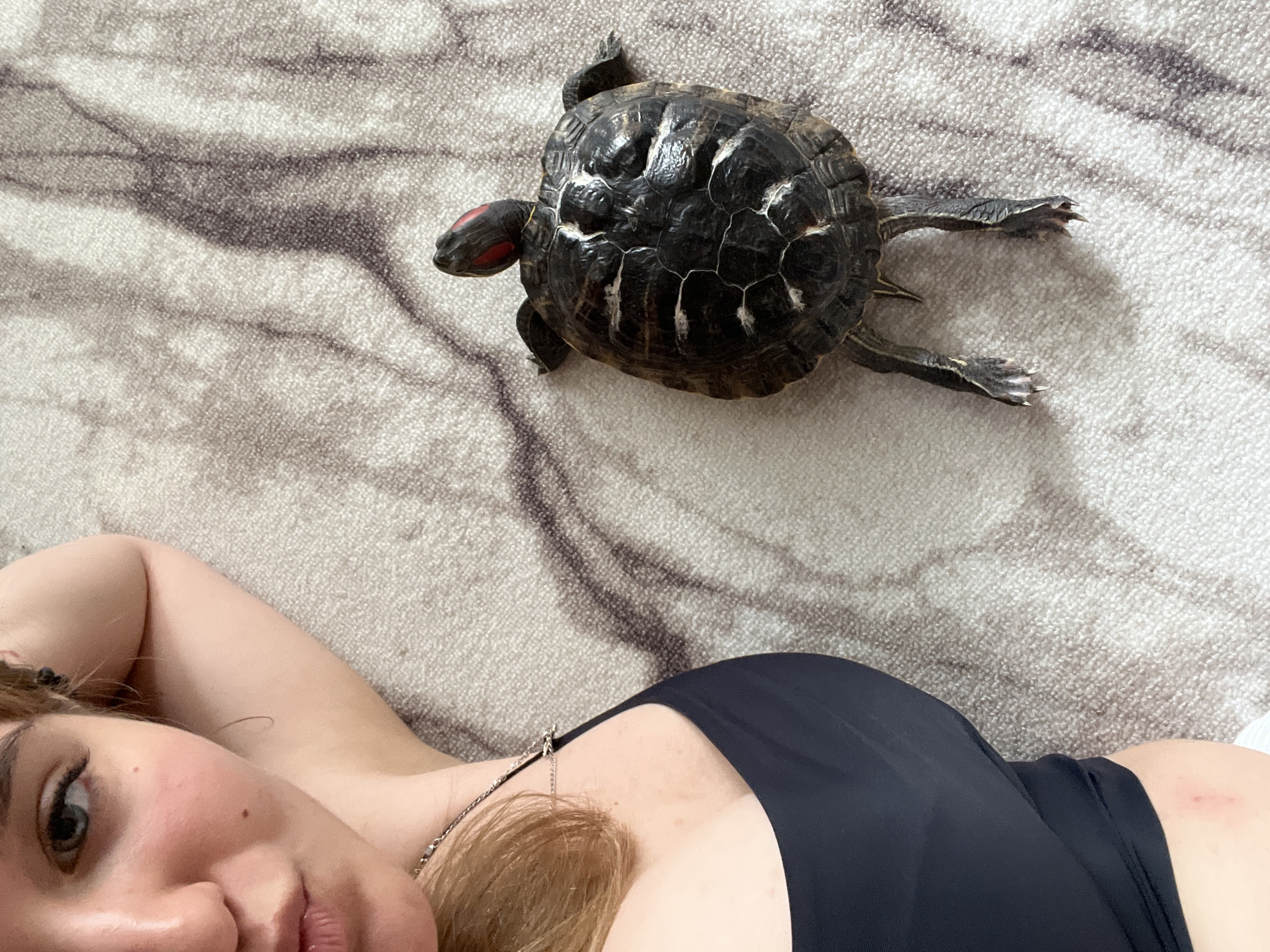 A close-up, overhead shot features a young woman lying on her back on a light-colored carpet with a marble-like pattern. Her head is turned towards the camera, with her left arm resting behind her head. She has blonde hair, light skin, and is wearing a black tank top and a silver necklace. To her right, a red-eared slider turtle is positioned on the carpet. The turtle is facing away from the camera, with its head extended and its legs splayed out. The image appears to have been taken indoors, possibly in a bedroom or living room. The location in Chaikovsky, Russia, is not discernible from the image itself.