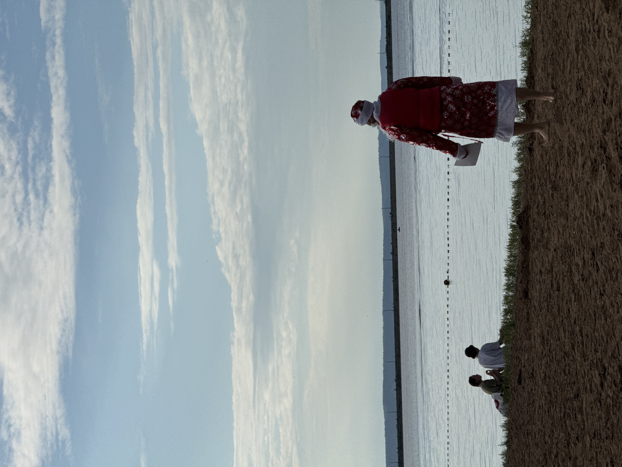 The image shows a group of people near a body of water, possibly a lake or sea, with a beach or embankment in the foreground. The sky is a pale blue with thin, wispy clouds. The sun appears to be high in the sky, suggesting it is daytime.
In the foreground, a person dressed in a Santa Claus costume, complete with a red suit and white trim, is standing. This person is holding a white object, possibly a letter or a gift. Further back, closer to the water, two other individuals are seated on the embankment. One is wearing a white garment, and the other is in a green shirt. They appear to be interacting with each other.
The water is calm and reflects the light from the sky. A line of buoys or markers stretches across the water in the distance. The embankment is covered in dry, sandy soil and some sparse green grass. The overall mood of the image is peaceful and serene. There is no visible text in the image.