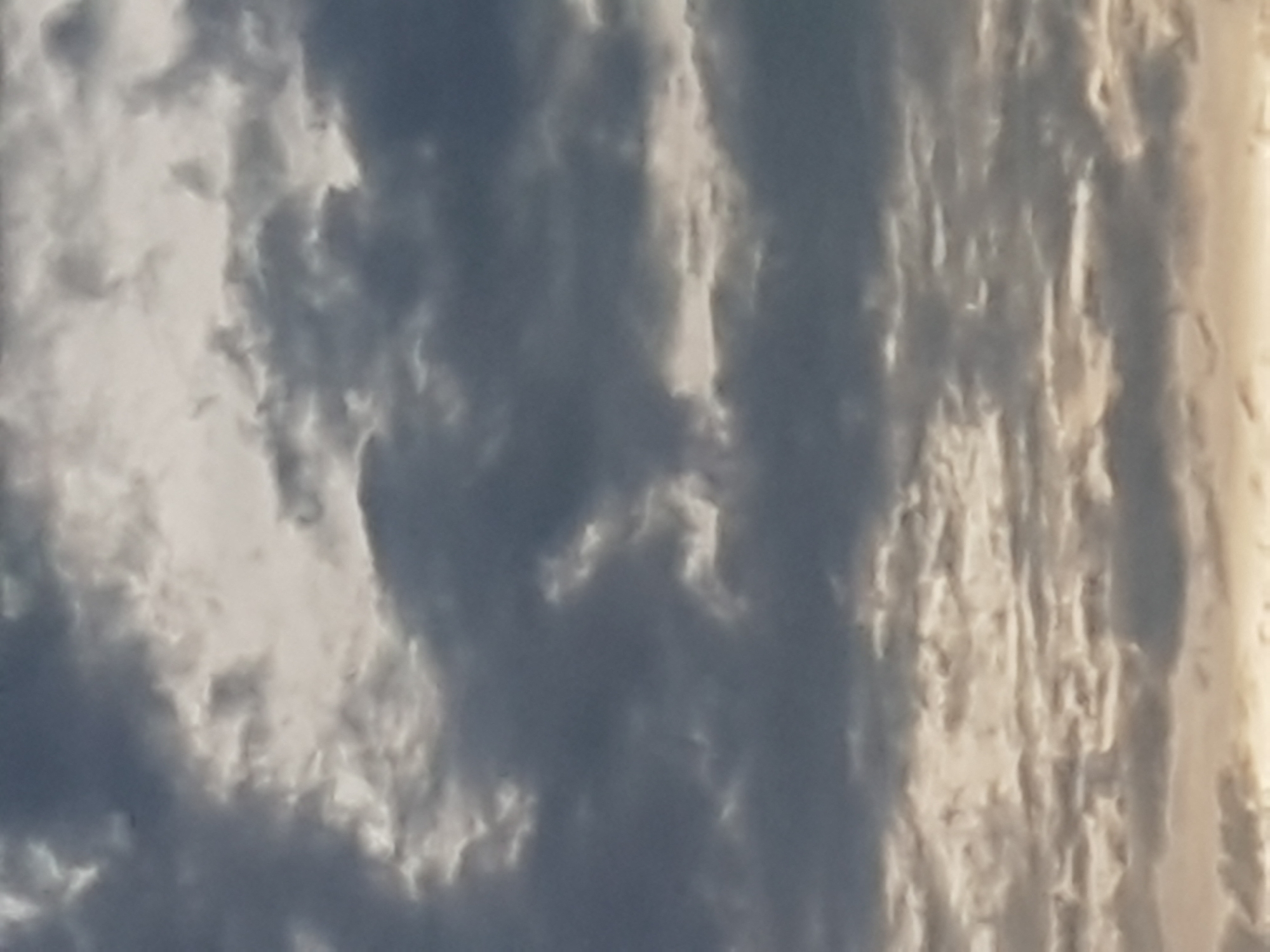 The image depicts a sky filled with dramatic, dark gray clouds with patches of bright light breaking through. The clouds are varied in texture and density, suggesting a dynamic weather pattern, possibly indicative of an approaching storm or the aftermath of one. The light source, likely the sun, is partially obscured, creating a diffused and somewhat ethereal illumination across the sky. The overall impression is one of a moody and overcast sky. There are no visible landmarks or specific environmental features to indicate a city location.