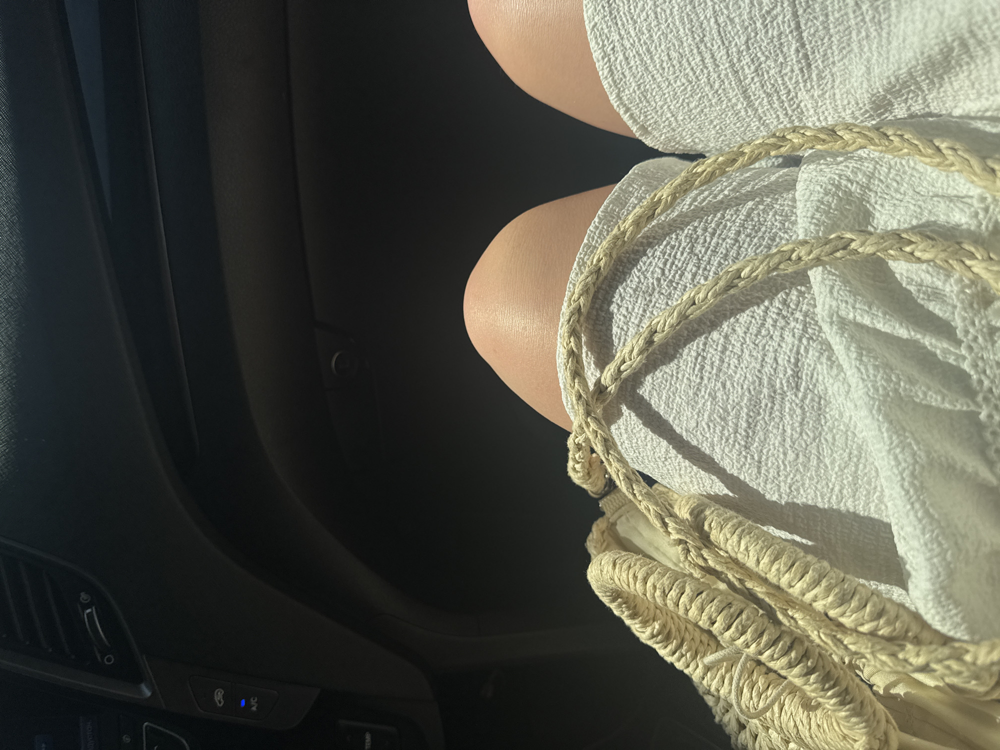 A view from inside a car shows the legs of a person wearing a white, textured dress or skirt. A braided, light-colored purse strap rests on the fabric. The car's interior is dark, with visible air vents and air conditioning controls. There are no discernible elements that indicate the location is Yevpatoriya, Ukraine.