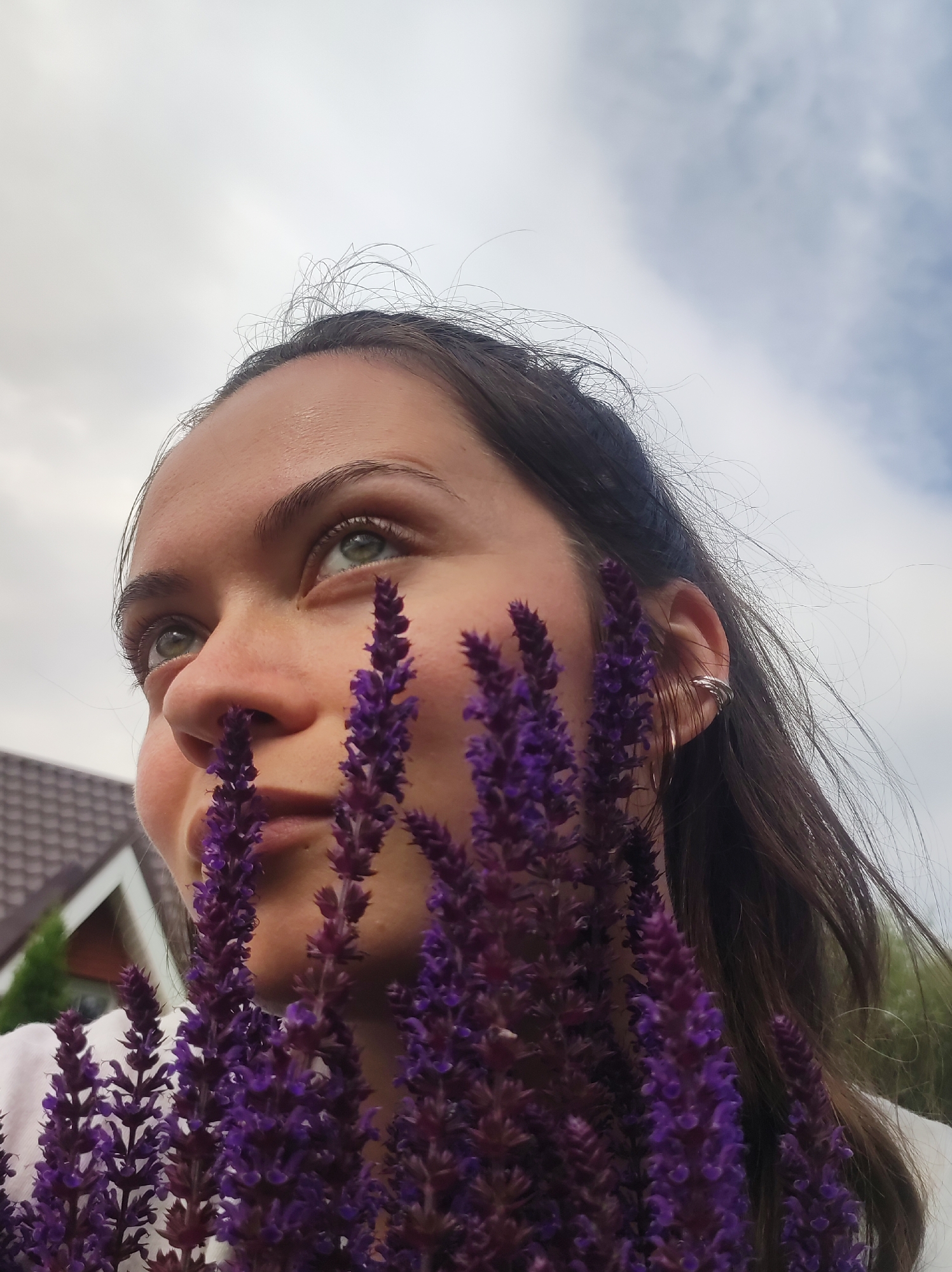 The image shows a close-up, low-angle shot of a young woman with dark hair and green eyes, looking upwards. She is partially obscured by several sprigs of purple flowers, which are in focus in the foreground. The woman is wearing a white top and a silver ear cuff. In the background, out of focus, is a section of a residential building with a brown tiled roof and some greenery. The sky above is overcast. The location is stated as Фоки, Russia, but no specific landmarks identifying this location are visible in the image.