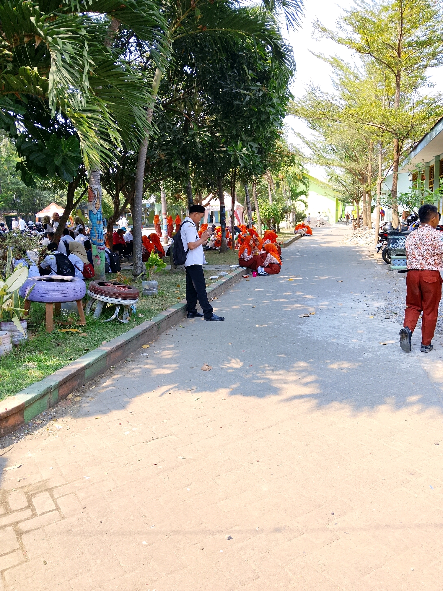 The image depicts a scene in front of a building, possibly a school, in Kramat, Indonesia. The foreground is dominated by a paved pathway with visible shadows from trees, indicating an outdoor setting under sunlight. A group of young people, many wearing orange headscarves and brown uniforms, are seated on the ground near the pathway. To the left, several individuals are sitting on repurposed tires that serve as makeshift seats, some appearing to be students. A man wearing a white shirt, dark pants, and a black cap is standing on the pathway, looking at his phone. Further down the pathway, another young person in a patterned shirt and brown pants is walking away from the camera. In the background, more trees line the pathway, and glimpses of buildings with green accents are visible. Motorcycles are parked in the background. The overall atmosphere suggests a casual gathering or break time.