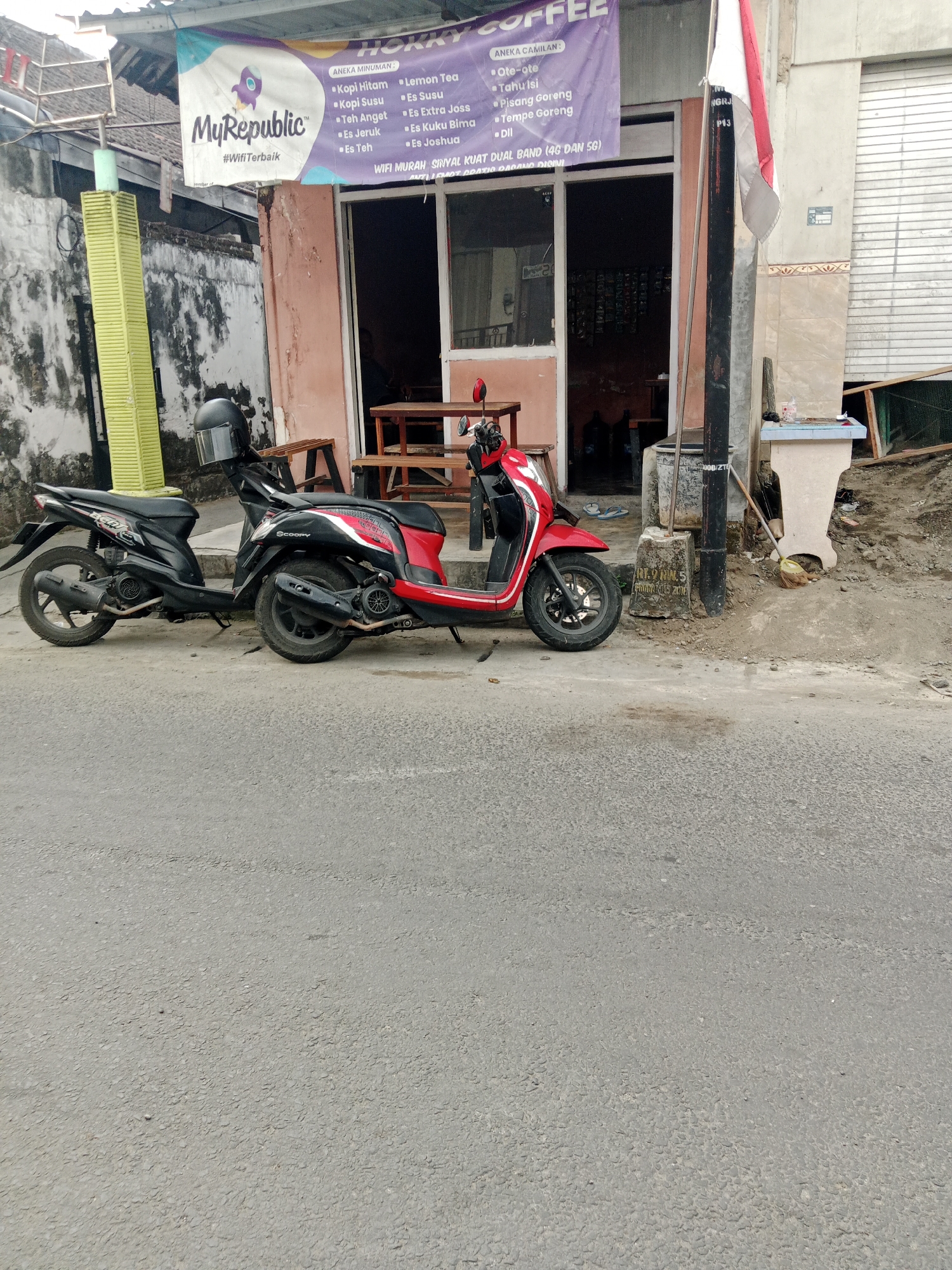 The image shows a street scene in Kediri City, Indonesia. Two scooters are parked on the side of the road in front of a building with a banner advertising "Hokky Coffee". The banner lists various drinks and snacks, including "Kopi Hitam", "Lemon Tea", "Ote-ote", and "Pisang Goreng". There is also text indicating "WIFI MURAH SINYAL KUAT DUAL BAND (4G DAN 5G)". The building appears to be a small shop or cafe. A red and white Indonesian flag is visible on a pole to the right of the building. The street is paved, and there is some debris on the ground near the building. The weather appears to be overcast.
