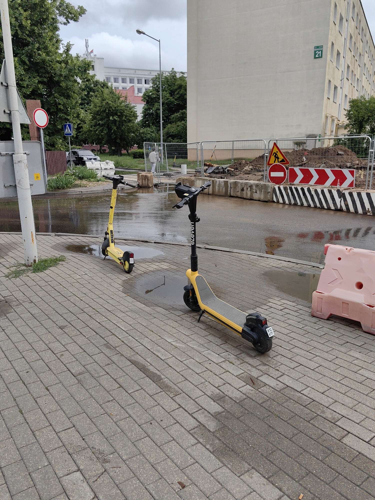 The image depicts a street scene with two yellow electric scooters parked on a paved sidewalk. The sidewalk is wet, suggesting recent rain. In the background, there is construction work, with barriers, piles of dirt, and traffic signs. A large building with windows is visible, and beyond that, trees and a cloudy sky. A street light is also present. The scene appears to be in a city, possibly Minsk, Belarus, given the presence of Cyrillic lettering on some signs and the architectural style of the building. One of the scooters has the word "eleven" written on its stem.