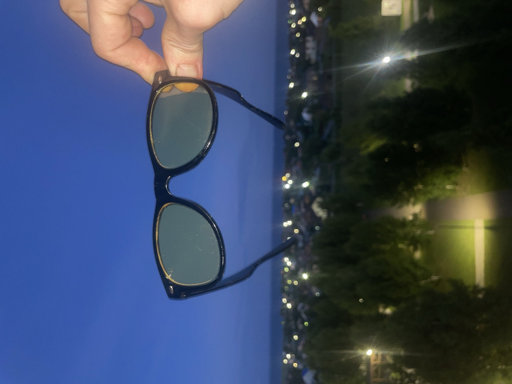 A person's hand holds a pair of black sunglasses with green lenses against a twilight sky. The sunglasses are positioned in the foreground, obscuring a blurred view of a cityscape in the background. The cityscape appears to be a residential area with houses and illuminated windows, suggesting it is dusk or early evening. Trees are visible in the midground, also appearing somewhat out of focus. The sky is a deep blue, transitioning to a lighter hue near the horizon. The overall impression is a casual moment, possibly capturing a view through sunglasses at sunset or after dark. The location is identified as Laishevo, Russia.