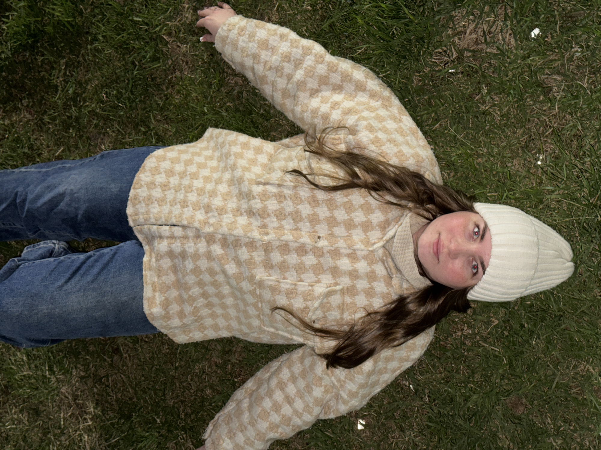 An overhead shot captures a young woman lying on her back on a patch of green grass. Her head is positioned towards the lower right of the frame, with her body extending upwards and to the left. She is dressed in a light-colored, possibly beige and off-white, patterned jacket with a small checkerboard or houndstooth design, worn over a lighter, possibly cream-colored top. Her legs are clad in blue jeans. A white, ribbed knit beanie covers her head, and her long, dark brown hair spills out from under it onto the grass and over her shoulder. Her arms are outstretched to her sides, resting on the grass. She looks directly up towards the camera with a neutral, calm expression. The lighting suggests it is daytime. There are no specific environmental cues in the image to indicate the location, but it is provided as Chaikovsky, Russia. No visible text is present.