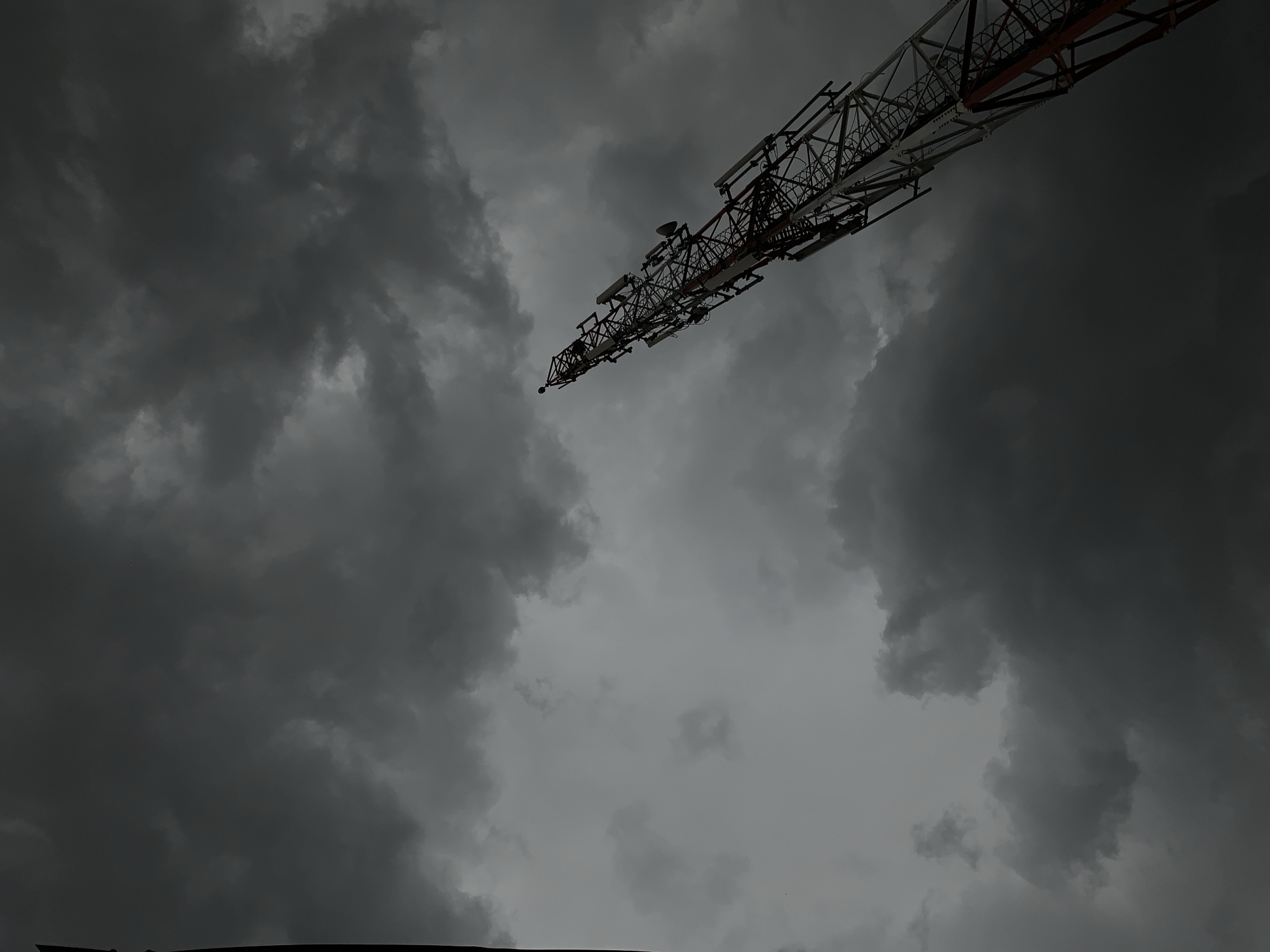 The image depicts a low-angle view of a communication tower against a backdrop of dark, stormy clouds. The tower is constructed from a lattice of metal beams and features multiple antennas and equipment boxes. The sky is overcast with dense, gray clouds obscuring any sunlight. The scene is located in the city of Rafin Par, Nigeria.