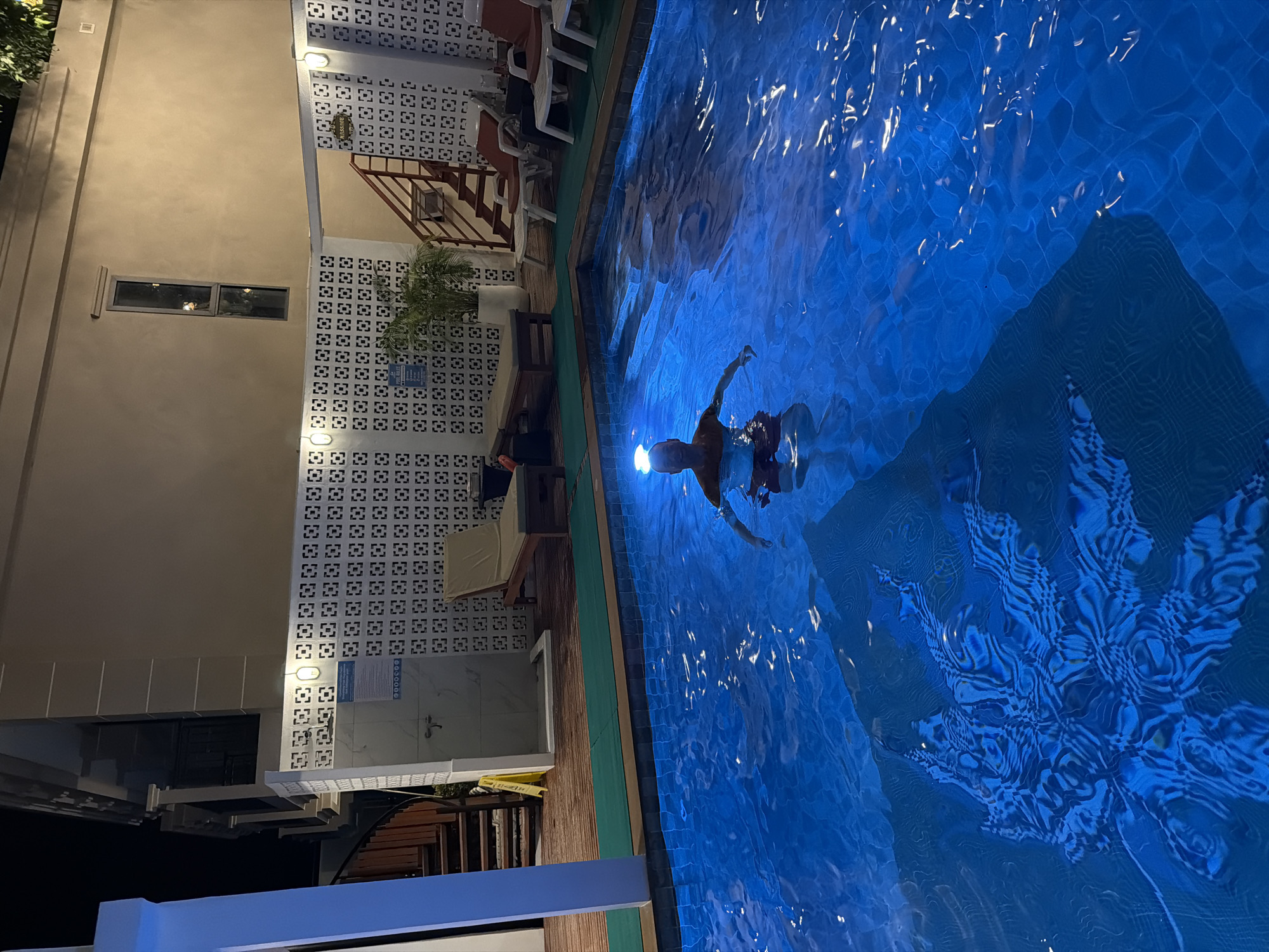 The image shows a nighttime scene at what appears to be a swimming pool in บ้านบางรักษ์, Thailand. A person is swimming in the blue-lit pool, with a bright light emanating from their head. The pool has a design at the bottom. Adjacent to the pool is a building, with visible architectural details including what appears to be decorative concrete blocks, windows, and stairs leading to an upper level. There are lounge chairs visible near the building. The scene is well-lit.