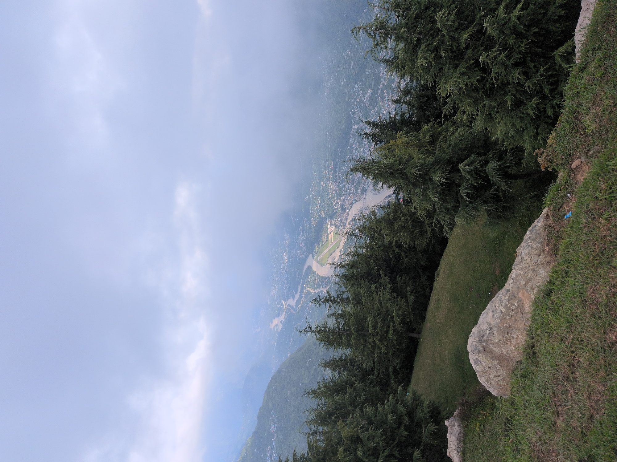 The image depicts a panoramic view of the city of Kullu, India, nestled within a valley. In the foreground, a grassy slope is dotted with large rocks, with a cluster of evergreen trees forming a natural barrier. Beyond the trees, the Beas River winds its way through the valley, bordered by a dense urban landscape. The city is characterized by its buildings, roads, and an airport runway, all partially obscured by a layer of mist and clouds that hang low in the sky, creating a somewhat ethereal atmosphere. The surrounding mountains are covered in lush greenery, with some areas exhibiting visible structures, suggesting terraced farming or settlements. The overall scene captures the juxtaposition of nature and human habitation in this picturesque Indian valley.