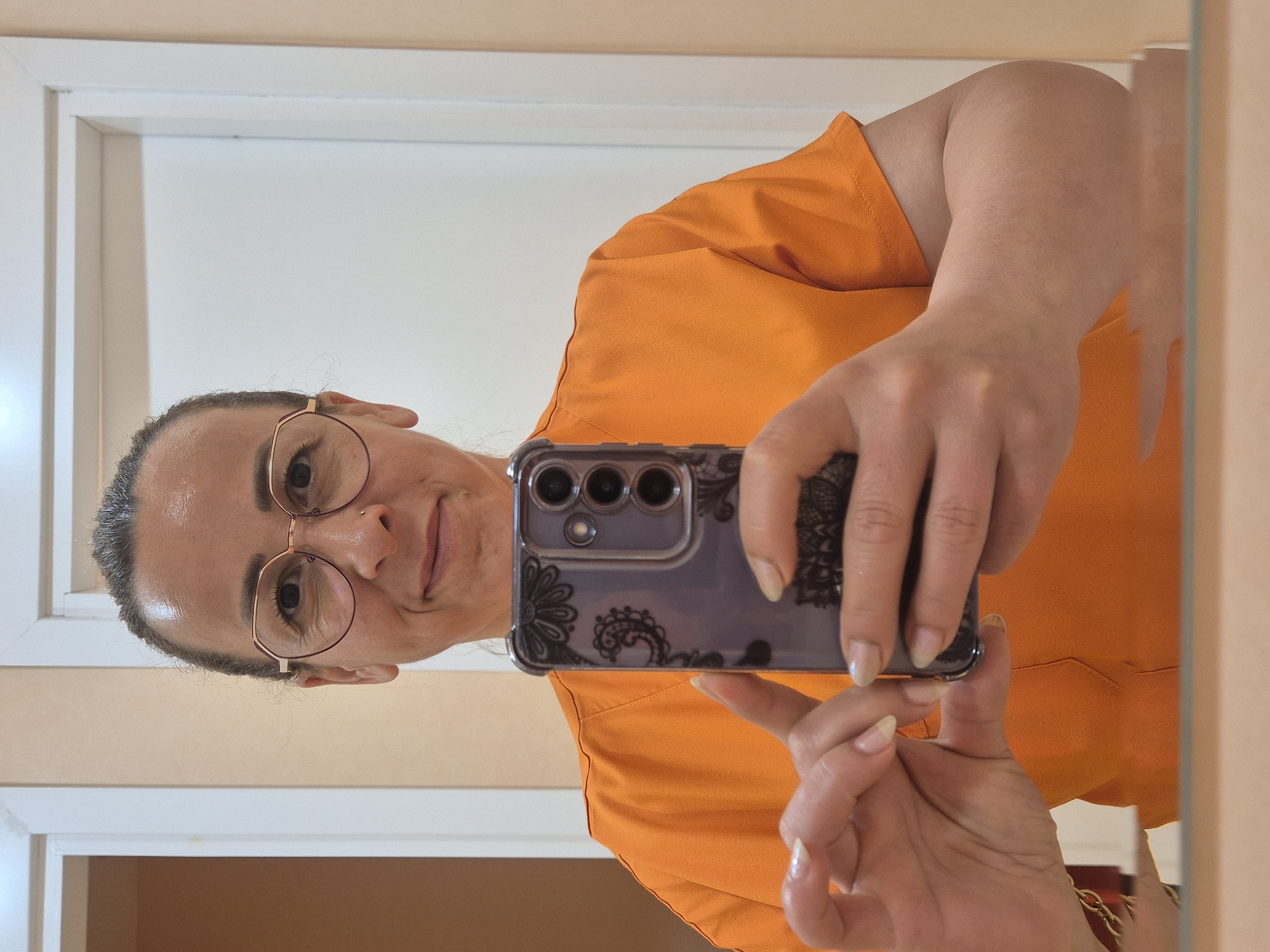 An adult individual is captured taking a mirror selfie, with the image originally rotated 90 degrees counter-clockwise. The person, appearing to be an adult female, is looking towards their reflection with a slight, pleasant smile. They have dark hair pulled back, revealing some visible grey strands, and are wearing round, gold-framed glasses. A small piercing is visible above their upper lip.
The individual is dressed in a bright orange, short-sleeved top, possibly a scrub top or a casual shirt. They are holding a dark-colored smartphone, equipped with a translucent case adorned with a black floral or paisley pattern. The phone's multi-lens camera array is clearly visible on its back. Both hands are holding the phone, with well-kept, naturally polished fingernails.
The background reflected in the mirror consists of plain, light-colored walls, likely beige or cream, and a white door frame or window frame, indicating an indoor setting. There is no visible text in the image.