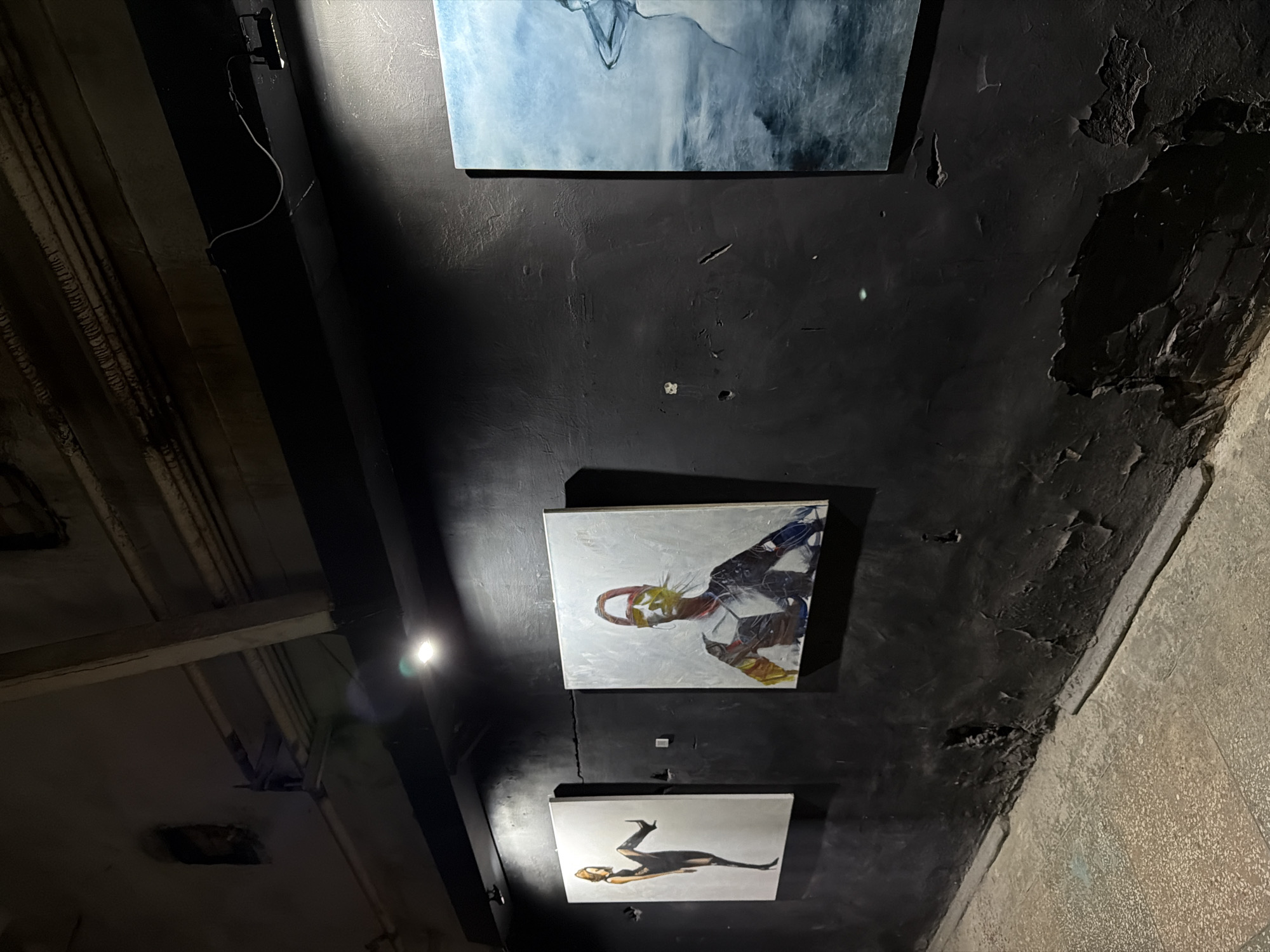 The image shows a view of the ceiling of a building or structure in Saratov, Russia, with three paintings or artworks mounted on the ceiling. The ceiling is painted black and appears to be in a state of disrepair, with peeling paint and rough surfaces. The paintings are square or rectangular in shape and depict abstract or stylized figures. The top painting is predominantly blue and white. The middle painting contains abstract figures, with colors like red, yellow, white, and blue. The bottom painting shows a dark figure with a lighter head and a tail or limb extending backwards. A light fixture or source is visible near the bottom left corner of the image. There are support beams across the ceiling. The right side of the frame shows the wall is peeling with a stone covering it.