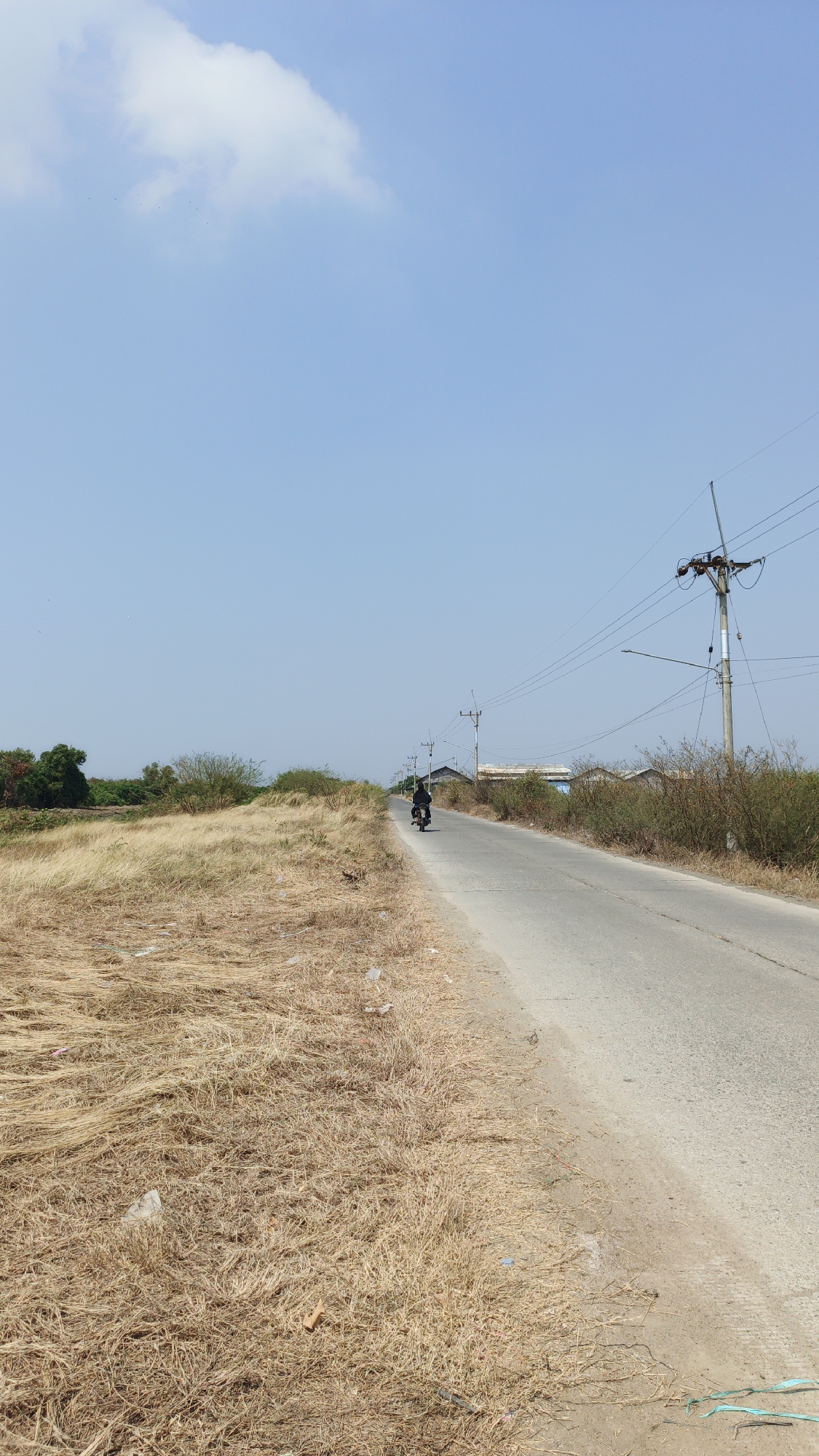 The image depicts a rural road in Krimun, Indonesia, on a clear, sunny day. The road, which is paved and appears to be in moderate condition, stretches into the distance. To the left of the road, a wide expanse of dry, golden grass dominates the landscape, with a few scattered bushes and trees visible further back. To the right of the road, there is a line of low-lying buildings, likely residential, behind a strip of sparse vegetation. Power lines and utility poles are present along the right side of the road, indicating some level of infrastructure. A lone individual on a motorcycle is traveling away from the camera on the road. The sky is a pale blue with a single, wispy cloud. The overall impression is one of a quiet, somewhat arid rural environment.