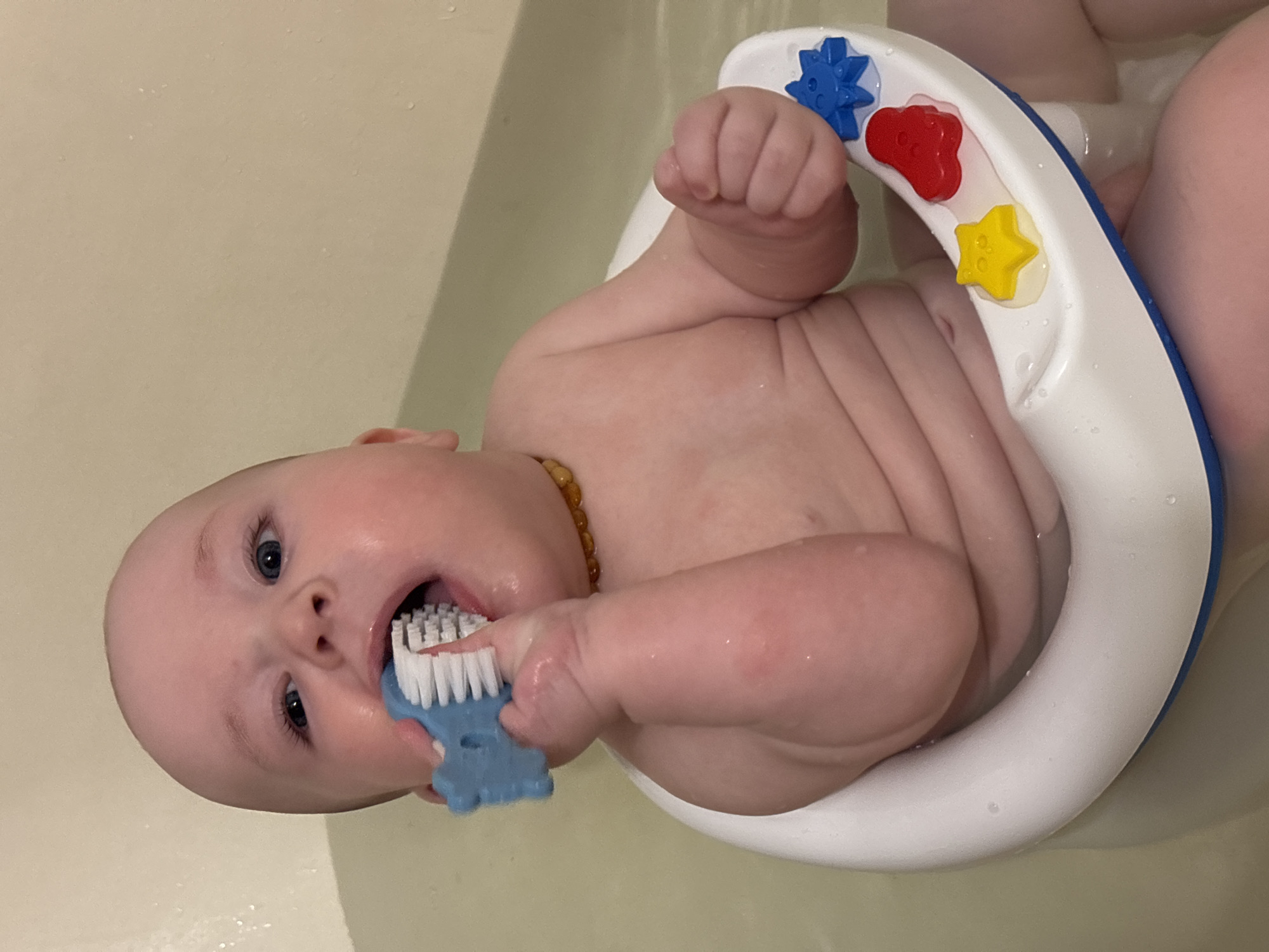 In a close-up, eye-level shot, a baby sits in a bathtub wearing a white and blue bath ring with colorful star and flower-shaped decorations. The baby, who appears to be teething, is chewing on a blue toothbrush. A beaded necklace is visible around the baby's neck. The bathtub is filled with water. The image was taken indoors. There is no indication or evidence that the image was taken in the city of Марковский, Russia.