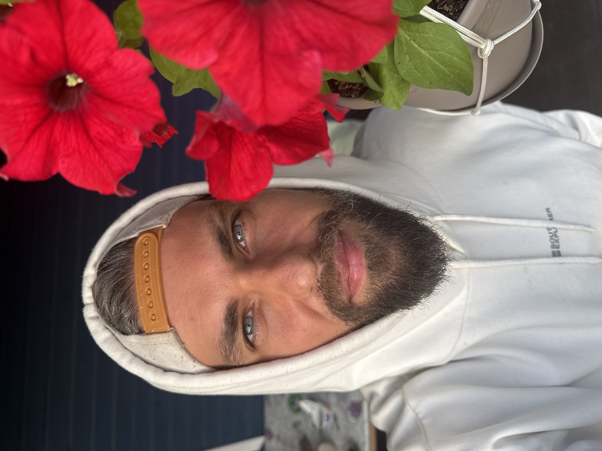 A man with blue eyes and a beard is photographed in close-up, wearing a white hoodie and a light-colored baseball cap. He is positioned to the left of the frame, with bright red petunias dominating the right side. The background appears to be a dark blue, weathered wooden surface, possibly a fence or wall. The man's gaze is directed towards the camera, and he is smiling slightly. The lighting suggests an outdoor environment, possibly during the daytime. There are no discernible landmarks or urban features to indicate a specific location within the city of Kuti, Russia. The image appears to be a selfie, given the perspective.