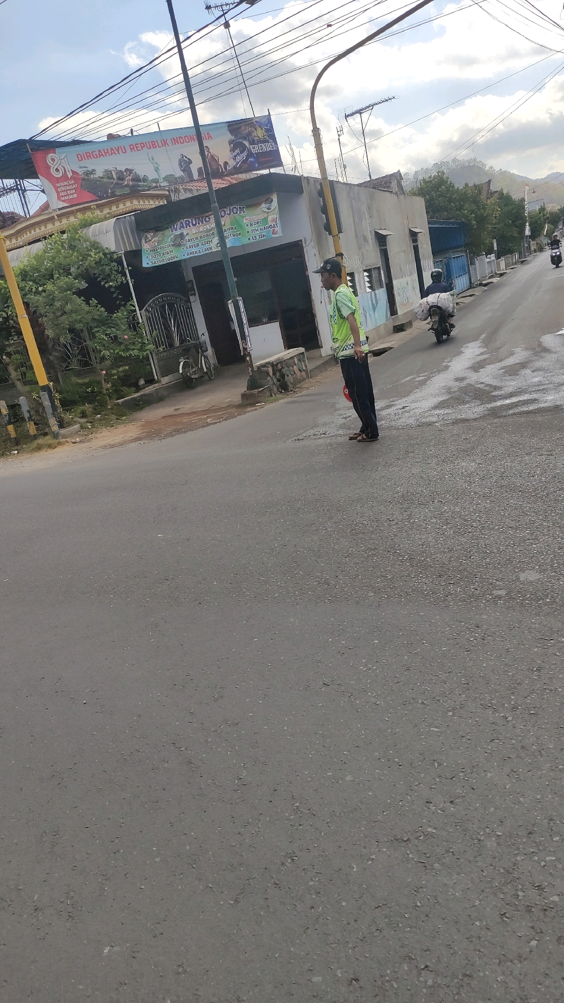 In the city of Ngrendeng, Indonesia, a man in a reflective vest directs traffic on a paved road. He stands on the right side of the road, facing left, wearing a dark cap and dark pants. His vest is bright green with white reflective stripes. Behind him, a motorcycle passes, with a rider and passenger visible. To the left of the road, a building displays a banner reading "DIRGAHAYU REPUBLIK INDONESIA," indicating a national celebration. The building also features signage for "WARUNG OJOK," suggesting it's a food and beverage establishment. Several power lines crisscross overhead, and trees line the background. The sky is partly cloudy.
