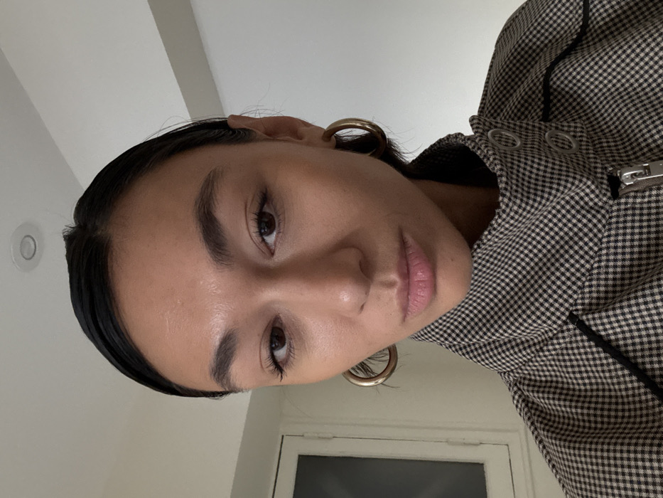 The image displays a close-up, chest-up portrait of a female subject, estimated to be in her 20s or 30s, with olive skin, dark hair pulled back, dark eyes, and full lips. She wears large gold hoop earrings. Her attire consists of a high-collared jacket or top with a small black and light-colored (beige or white) check pattern, suggestive of houndstooth or gingham. The collar features two visible silver-toned snap closures, and a silver-toned zipper pull is partially visible on the garment, which also includes black piping details. The subject maintains direct eye contact with the camera, her expression neutral with slightly pursed lips. The background reveals an interior space with plain light-colored walls and a ceiling containing a circular recessed light fixture. A partial view of a white-framed window or door is visible on the left side of the frame. The specific location within Paris, France, is not identifiable from the visual evidence presented.
