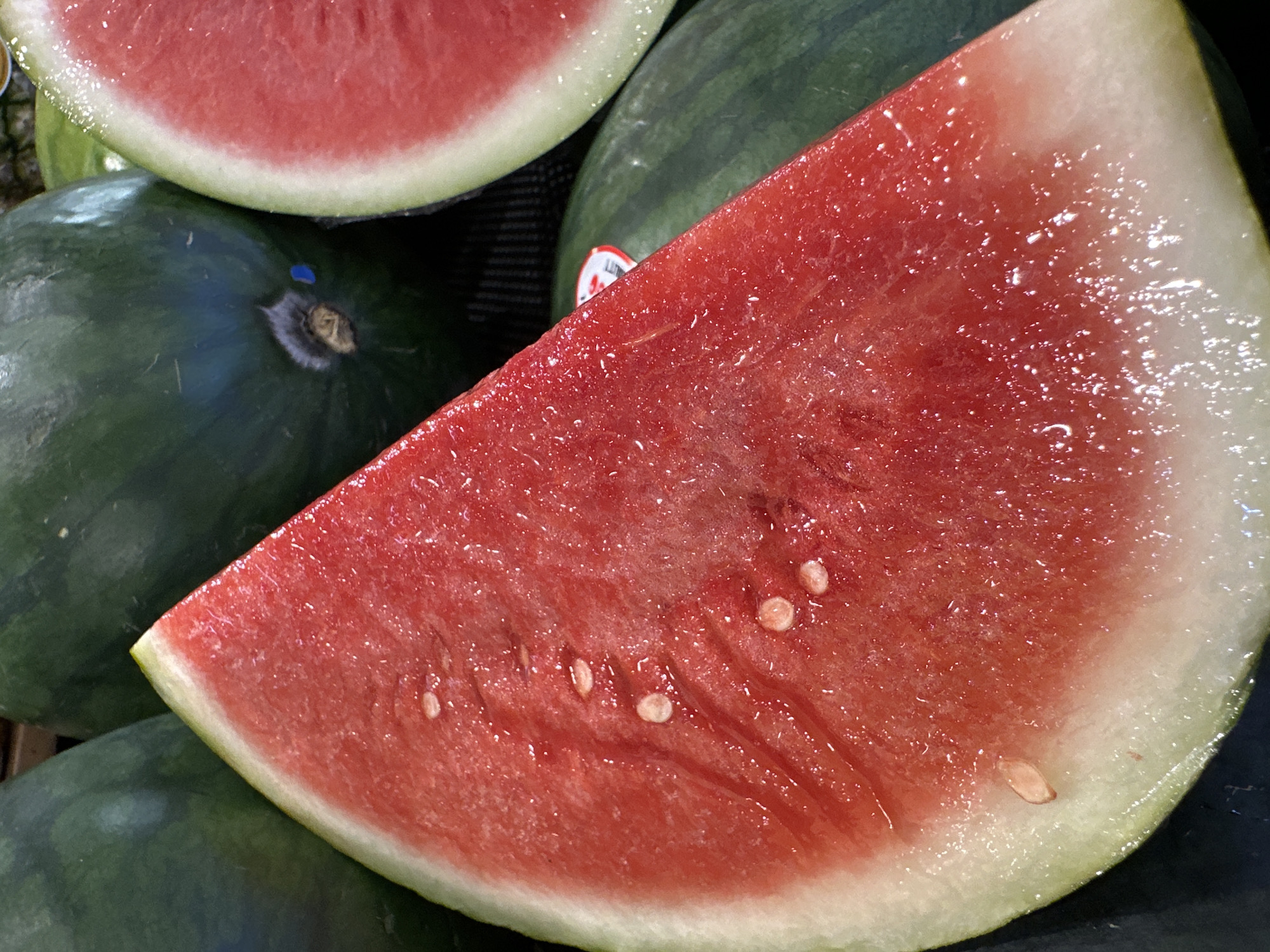 The image shows a close-up of a sliced watermelon, with the bright red flesh and white rind clearly visible. Several watermelon seeds are scattered throughout the flesh. In the background, whole watermelons with dark green rinds are visible. The setting appears to be a market or grocery store, as suggested by the way the fruits are displayed and the presence of a label on one of the whole watermelons. The lighting is bright, highlighting the texture and juiciness of the watermelon. There are no people or visible interactions in the image. The location of Kuta Selatan, Indonesia, is not discernable from the image itself.