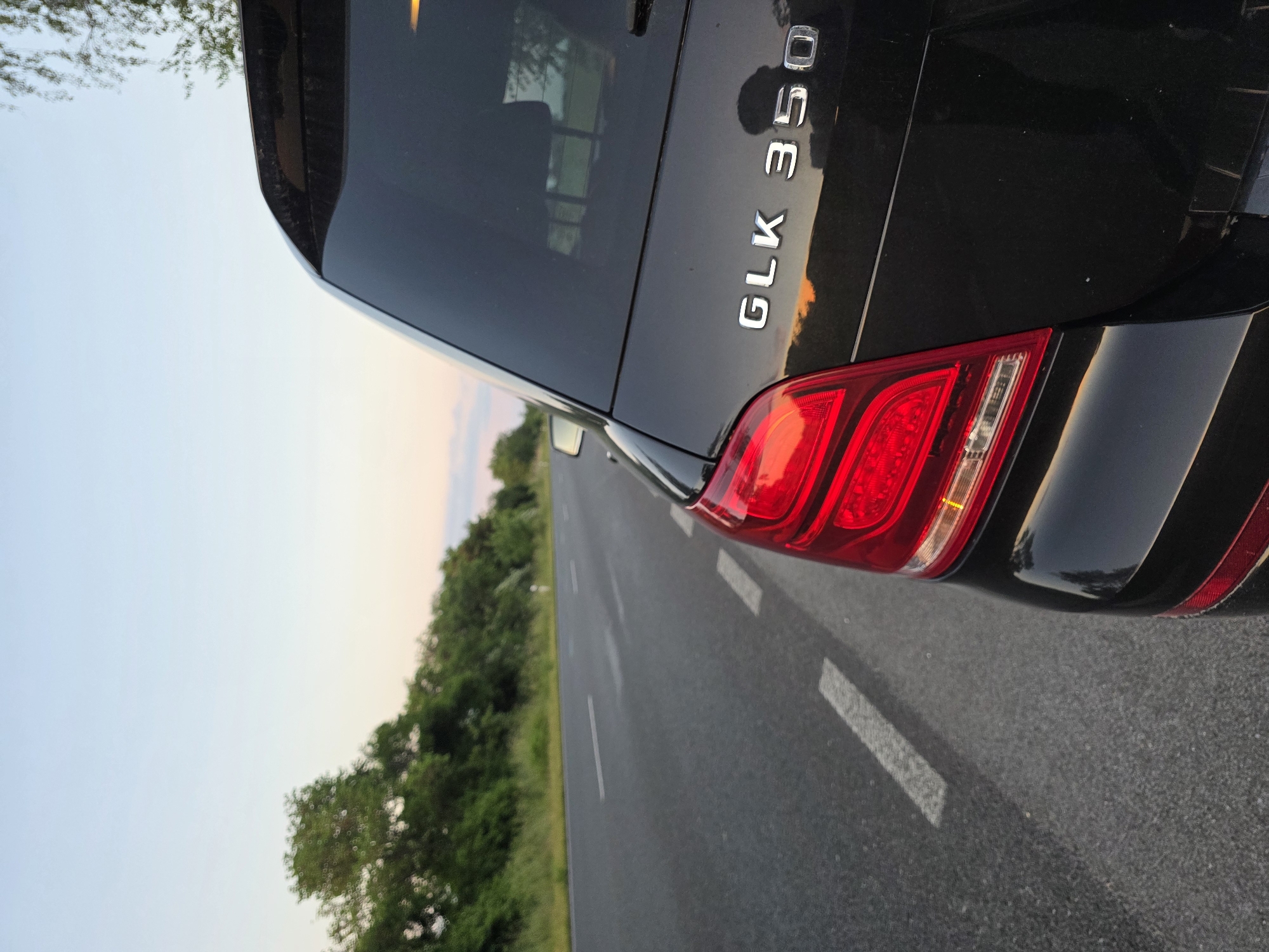 A black Mercedes-Benz GLK 350 is positioned on a two-lane road in an outdoor setting. The rear passenger-side corner of the vehicle is in the foreground, showing the tail light, GLK 350 badging, and part of the rear window. The road is marked with white lines, and a line of trees is visible along the side of the road. The sky has a gradient from blue to shades of orange and pink, suggesting either dawn or dusk. The precise location in Cheshnegirovo, Bulgaria, cannot be determined from the image, however, the environment appears to be a rural road.