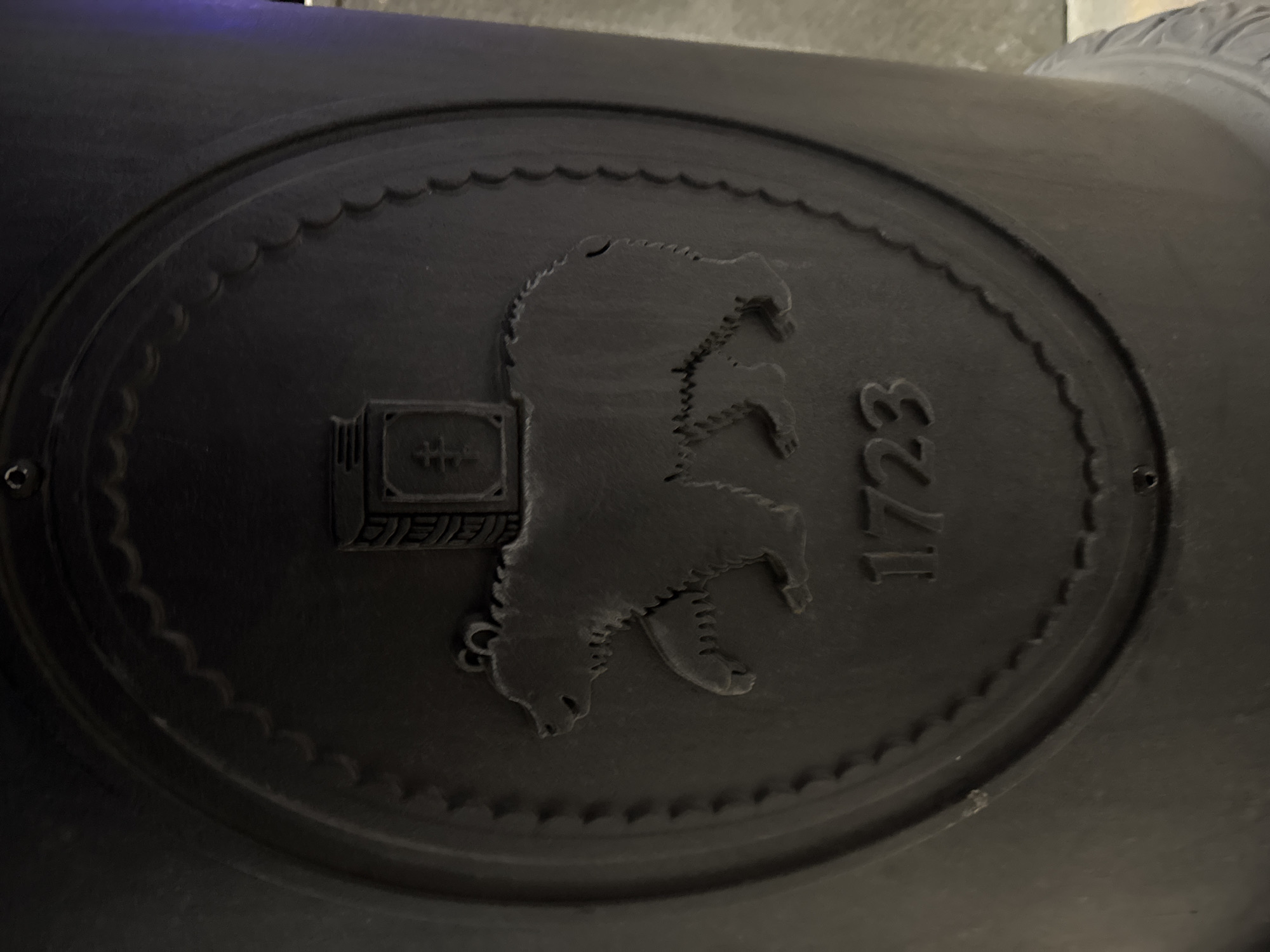 The image shows a dark, circular, embossed emblem. Inside the circle is a bear standing on its hind legs, facing right. To the left of the bear is a rectangular shape containing symbols. To the right of the bear is the number "1723." The outer edge of the circle has a scalloped pattern. The emblem is likely located in Perm, Russia, due to the presence of the bear, a symbol associated with the city.