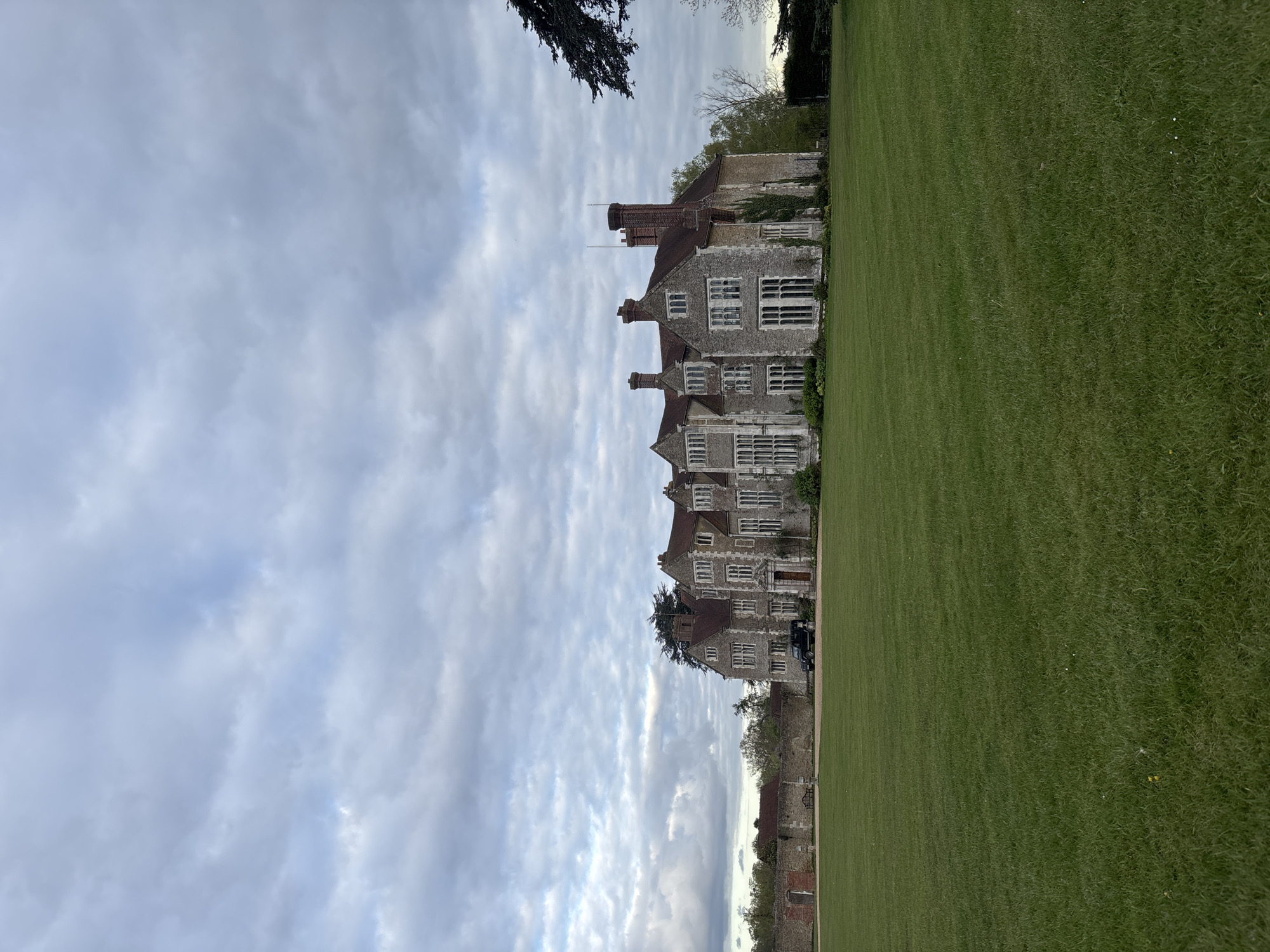 The image shows a view of Loseley Park, located near Guildford, United Kingdom. The large, stone manor house is multi-storied with numerous windows and chimneys. A sprawling, well-manicured green lawn stretches out in front of the house. Part of a brick wall is visible, running along the right side of the lawn. The sky is overcast with heavy cloud cover. A section of tree branches appears in the upper left corner of the image. No people are visible in the frame.