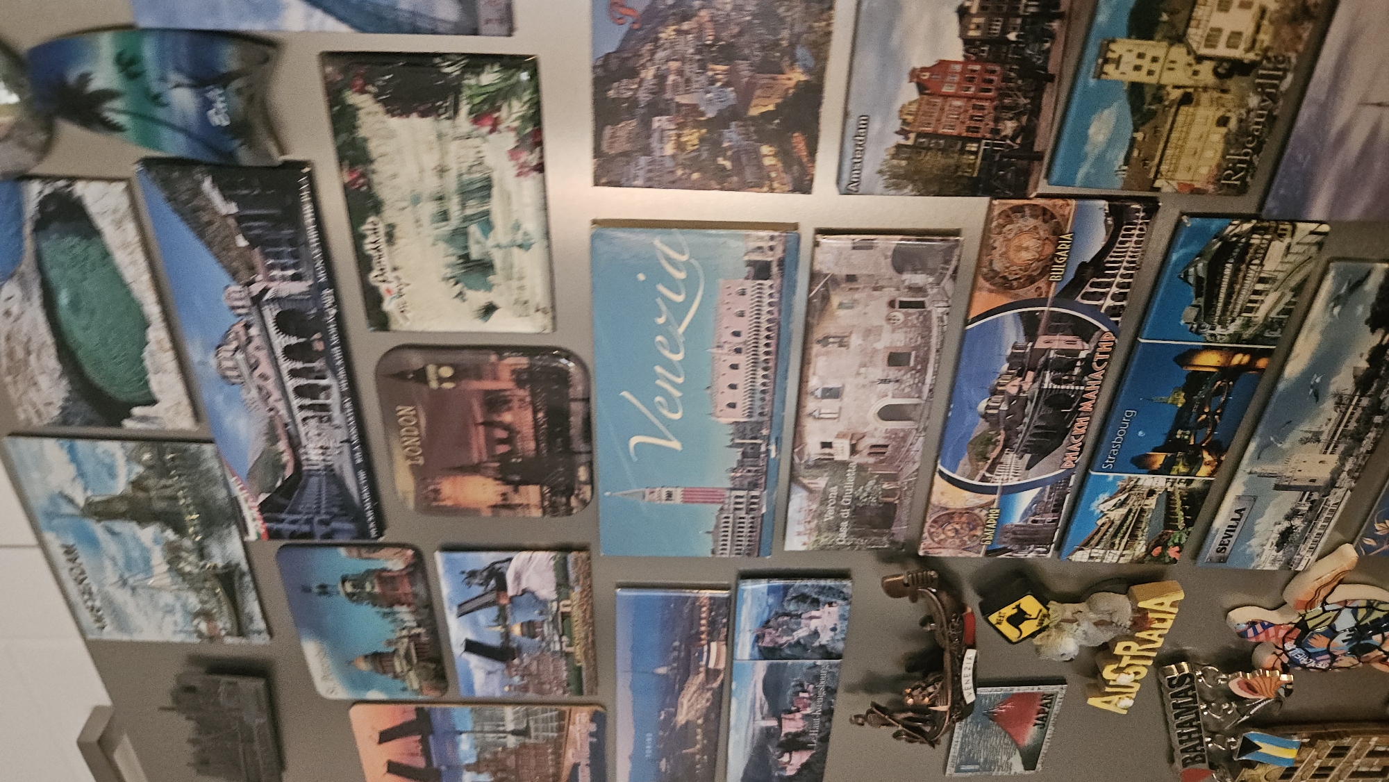 The image shows a collection of travel souvenir magnets on a refrigerator. Several magnets are rectangular with images of cities like "Venezia", "Amsterdam", "Ribeauville", "SEVILLA", "LONDON", and "Strasbourg". Other magnets represent countries such as "BULGARIA", "JAPAN", and "AUSTRALIA", along with destinations like "BAHAMAS" and "TEYCHELLES". One magnet appears to depict the "PHACKИ МАНАСТИР" which is most likely Rila Monastery in Bulgaria. There is no specific mention of Plovdiv, Bulgaria, in the visual data provided. A magnet labeled "Verona Casa di Giulietta" can also be seen. There are also some decorative magnets such as a turtle.