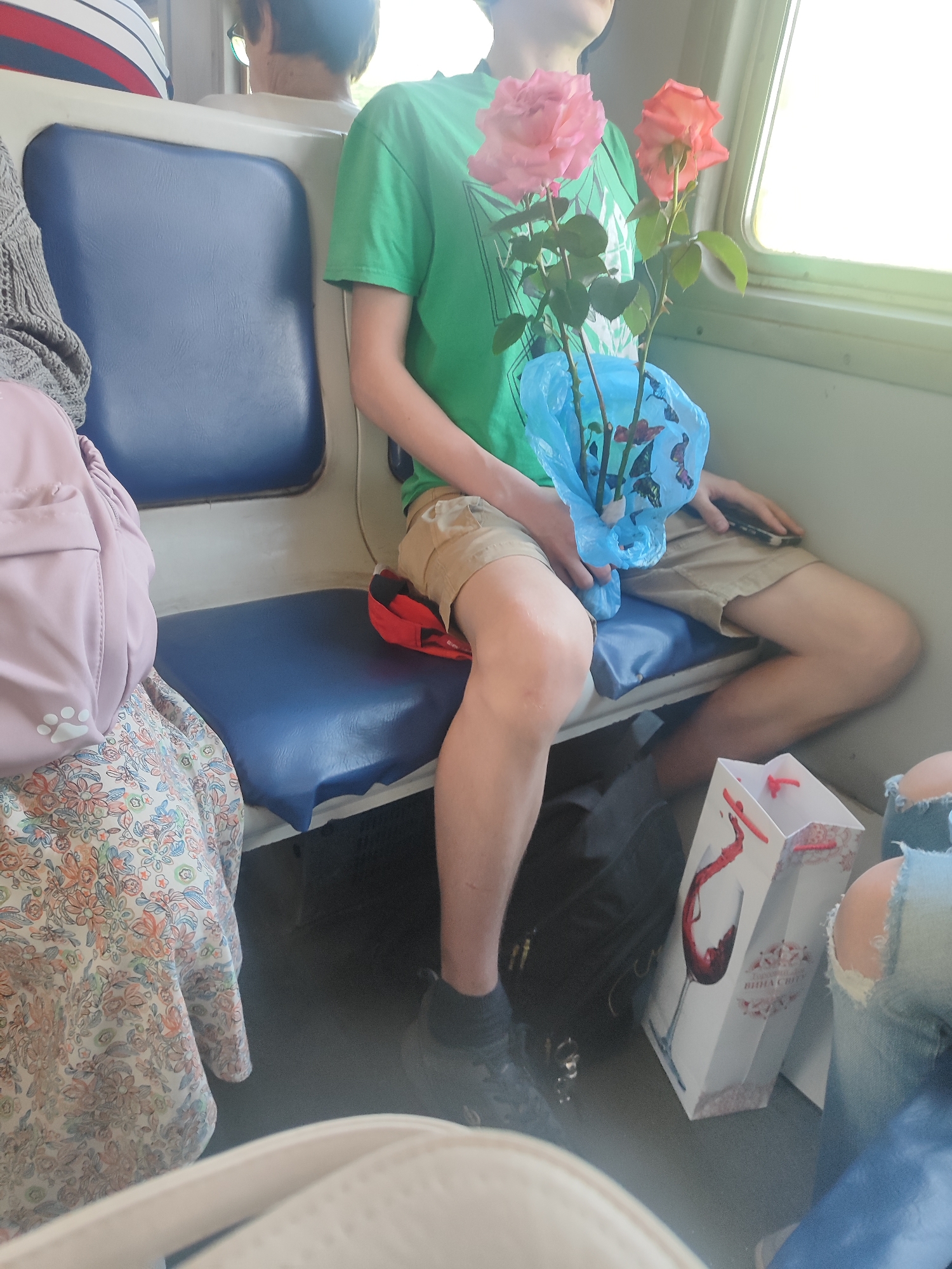The image depicts a medium shot of individuals seated on a public transport vehicle, likely a train, given the presence of luggage and the interior design. The scene is brightly lit, suggesting daytime.
In the foreground, a person is seated with two roses wrapped in blue plastic, held in their lap. One rose is light pink, and the other is a peachy orange. The person is wearing a green t-shirt and light brown shorts. Their legs are crossed, and they are holding a mobile phone in their right hand, which is resting on their right thigh.
To the left of this individual, another person is partially visible, wearing a light-colored patterned dress and a grey knitted garment. A light pink backpack with a paw print logo is placed on their lap.
In the lower right quadrant of the image, a white gift bag with a red wine glass and grapes design is visible, along with a section of denim jeans with rips, indicating another person is present. The floor of the vehicle is dark and appears to be utilitarian.
The seats are upholstered in blue, with a white frame. A window is visible to the right, letting in natural light. The overall impression is that of a casual journey.