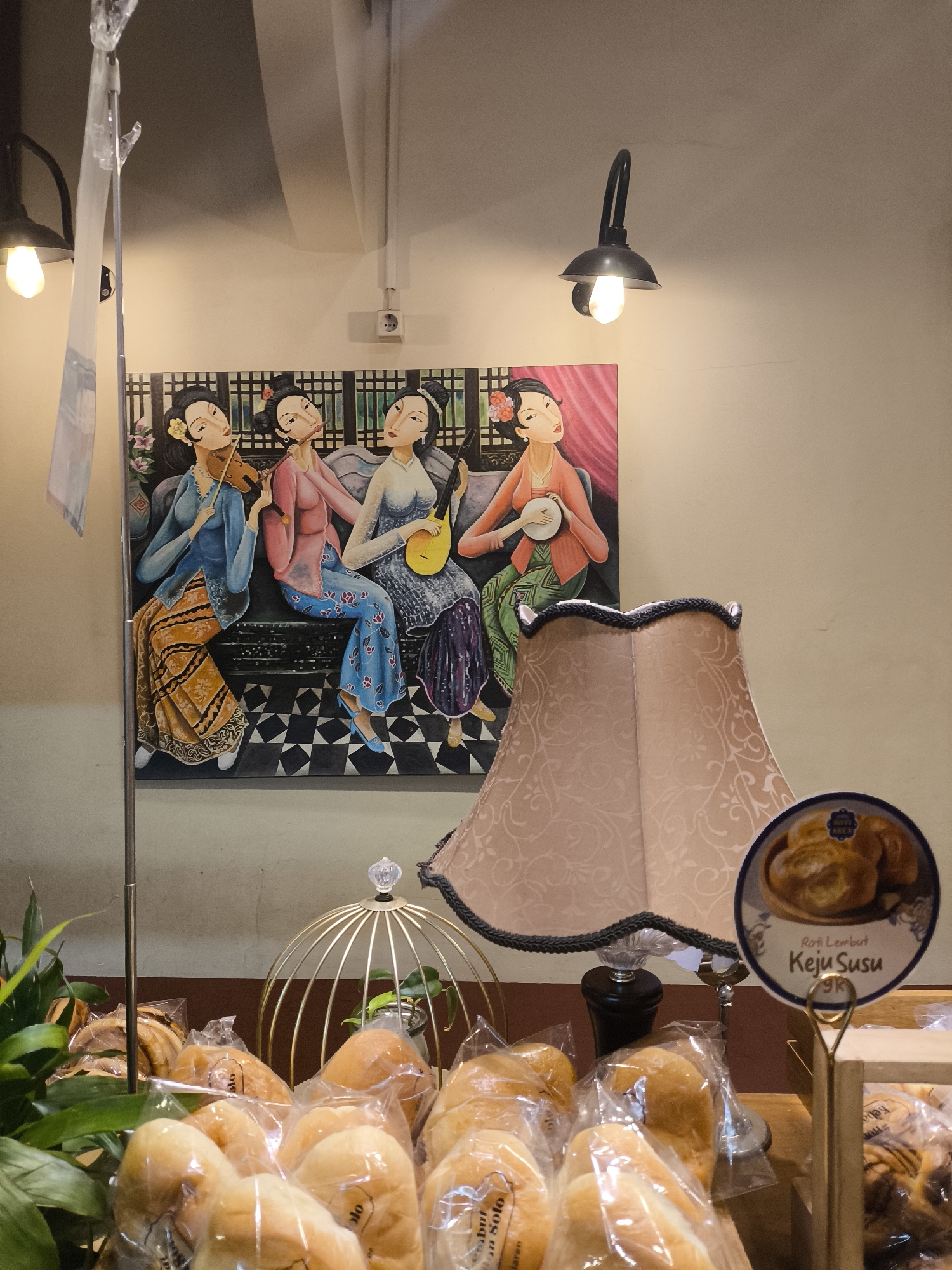 This image depicts a street-level view inside a bakery in Surakarta, Indonesia. The foreground is filled with an assortment of bread products, mostly enclosed in clear plastic packaging. The bread rolls appear soft and light-colored. Behind the bread, a decorative gold birdcage-like structure with a clear finial sits on a wooden surface. Further back, a vintage-style table lamp with a patterned pink lampshade is visible. To the right of the lamp, a circular sign displays an image of bread rolls and text in Indonesian that reads "Roti Lembut Keju Susu 9K," indicating soft cheese milk bread priced at 9,000 Rupiah. Above the lamp, a painting is hung on the wall. This artwork features four women in traditional Indonesian attire, seated and playing musical instruments. The background wall is painted a neutral beige color, and industrial-style light fixtures are mounted on the ceiling. The overall ambiance suggests a cozy and traditional Indonesian bakery.