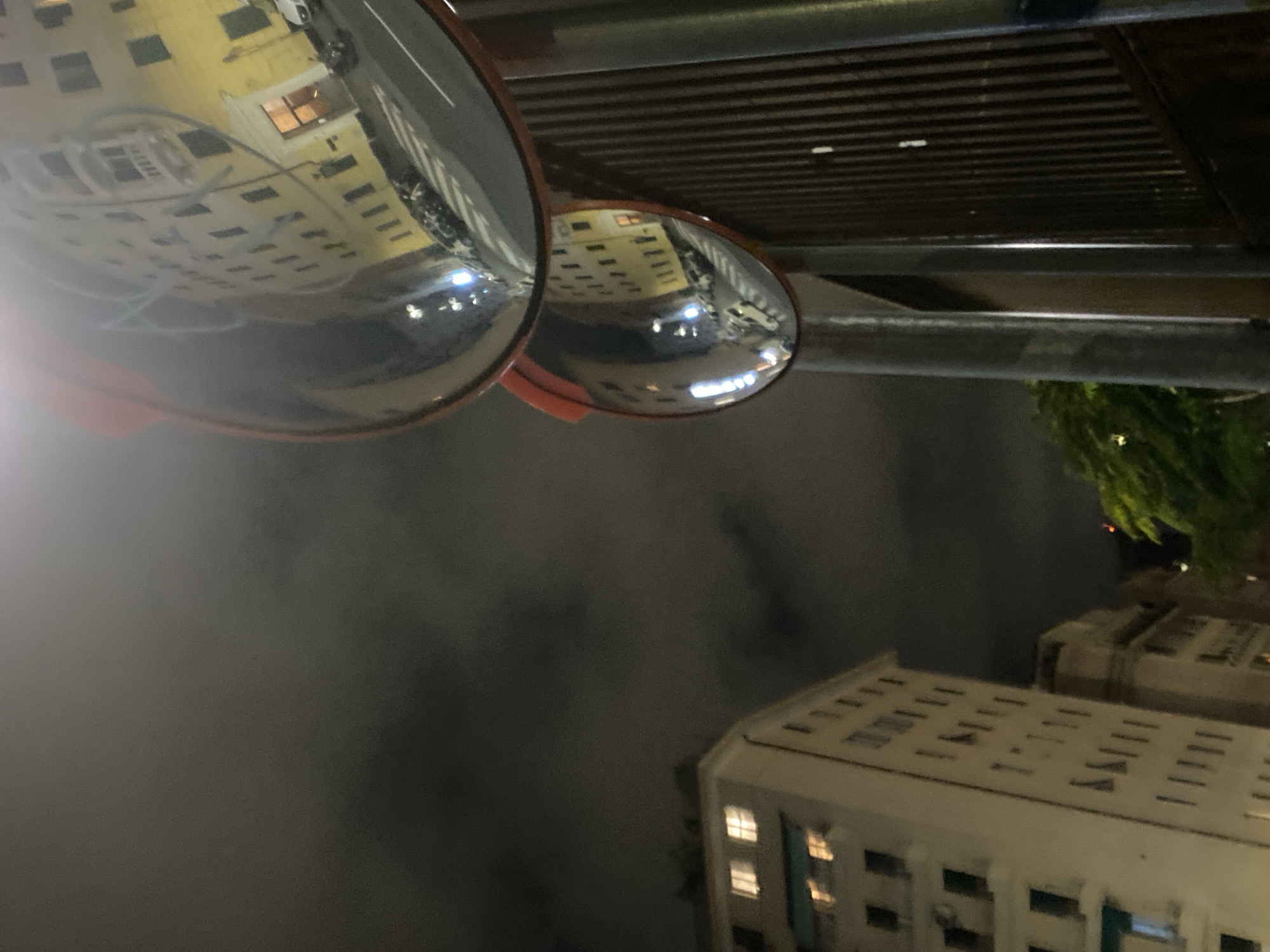 This image depicts a street scene at night in Genoa, Italy. Two convex mirrors are mounted on a pole, reflecting the surrounding environment. The larger, upper mirror shows a yellow building with multiple windows, a street with parked cars, and a pedestrian crossing. The smaller, lower mirror reflects a similar view of the yellow building and street, but from a slightly different angle. To the left of the mirrors, a white building with illuminated windows is visible. Further to the left and in the background, another building and some greenery can be seen. The sky is dark and cloudy, suggesting nighttime. The overall impression is an urban environment captured at night with the distorted reflections of buildings and street elements.