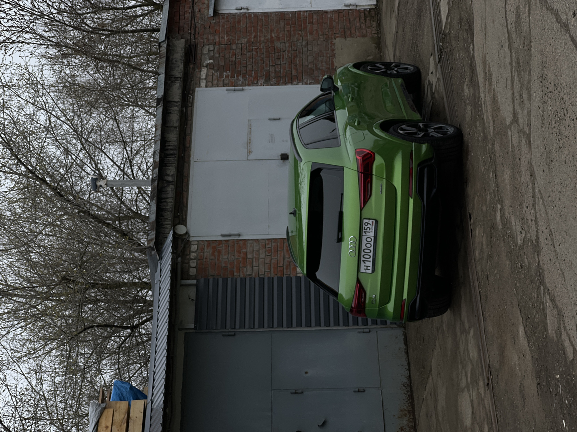 An image captures a bright green Audi SUV, likely a Q3 model, parked on a paved surface next to an older building in what appears to be an industrial or utility area in Chaikovsky, Russia. The car is positioned with its rear visible, featuring a license plate that reads "H100OO 159 RUSSIA" and the distinctive four-rings Audi logo.
The building to the left of the vehicle consists of a brick upper section and several large, grey, industrial-style garage doors or metal-clad panels below. Some corrugated metal sheeting is also visible on the building's facade. The paved ground shows signs of wear and possibly some dampness.
In the background, numerous bare tree branches stretch across the grey, overcast sky, suggesting a colder season such as late autumn, winter, or early spring. On the far left, partially obscured by the trees, there are stacks of wooden planks covered by a blue tarp, hinting at storage or construction activity nearby. A small, round, white object, possibly a security camera or light fixture, is mounted on the building's brick wall. No people are visible in the scene, and no specific activity is taking place beyond the car being parked. The overall impression is that of a quiet, somewhat utilitarian daytime setting under a cloudy sky.