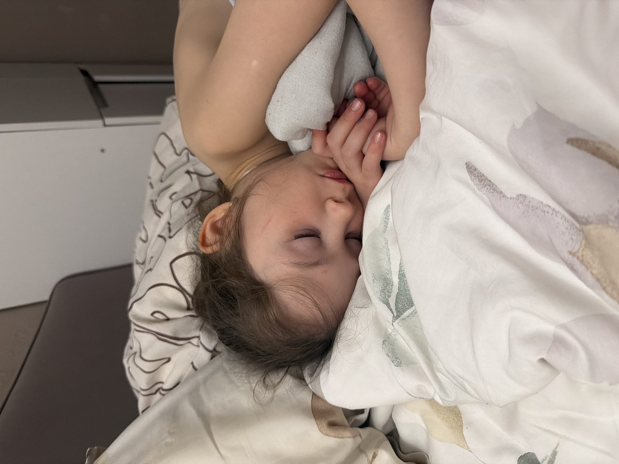 A close-up shot depicts a child sleeping. The child's face is partially obscured by their hands and the bedding. The bedding appears to be patterned with floral designs. The location is unclear, and no specific details suggest the city of Perm, Russia. Other objects visible include a dresser or bedside table partially visible in the background.